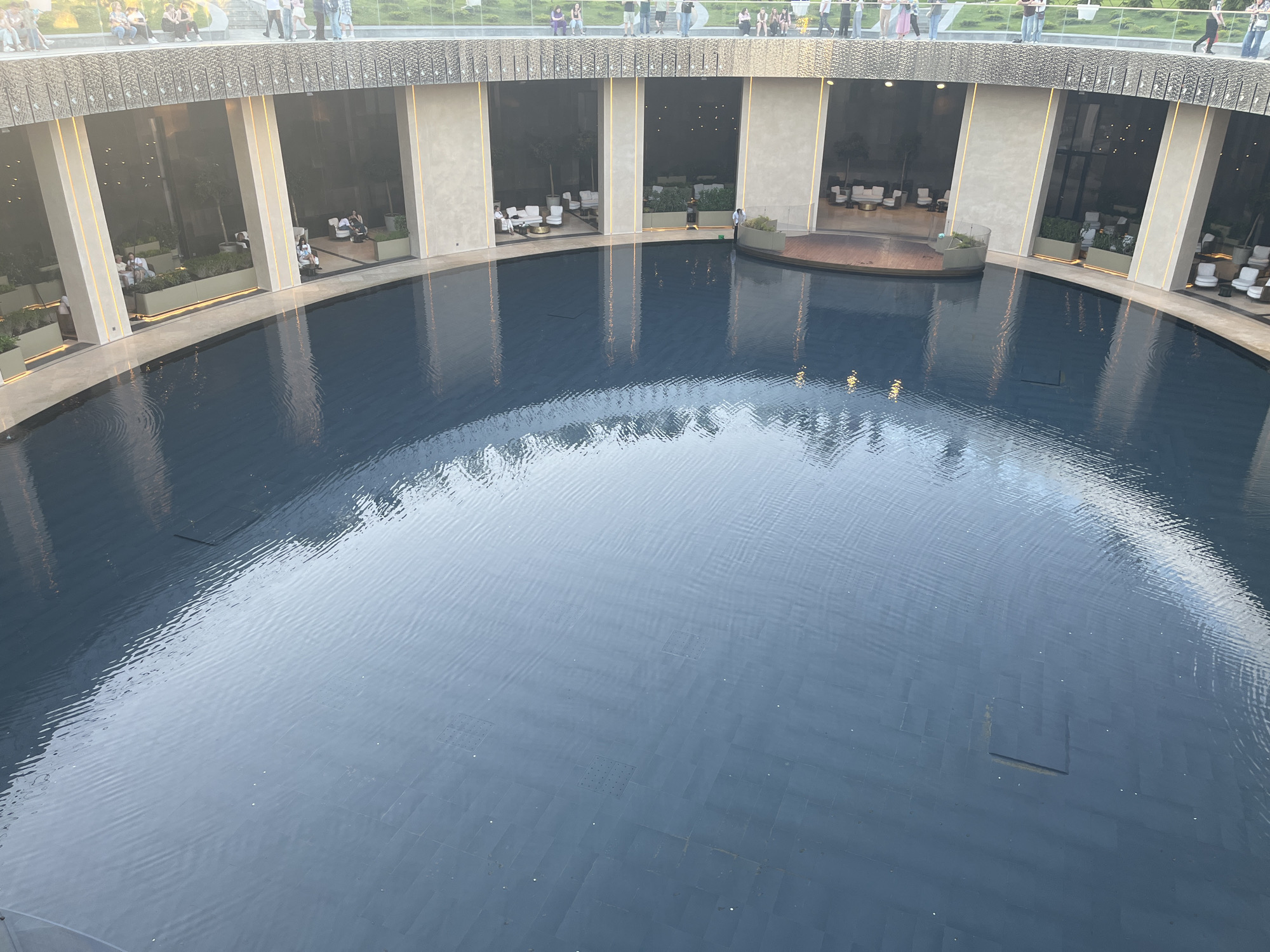 This media file presents an elevated view of a large, circular architectural space centered around a deep, dark water basin. The setting appears to be a modern, public, or commercial leisure area.
An upper-level promenade or walkway circles the entire structure, with numerous people visible, casually dressed, walking or standing and observing the scene below. This level features a textured, possibly metallic or stone, balustrade.
Below the promenade, a lower, covered level forms a continuous ring around the water. This area is defined by large, light-colored concrete pillars that alternate with expansive glass panels. Inside these glazed sections, various lounge-style seating arrangements are visible, featuring white sofas, armchairs, and tables, alongside planters with greenery. A few individuals are seated, relaxing in these areas. One person, dressed in a white shirt and dark trousers, stands by the water's edge on this lower level.
The prominent feature is the large, circular water basin in the foreground and midground. The water's surface, a dark blue-grey, exhibits gentle ripples and reflects the surrounding architecture and the sky above. The bottom of the basin, which appears tiled, shows square grid patterns in some areas. On the right side, a curved wooden platform extends slightly into the water, suggesting a unique viewing spot or a small stage.
The lighting is soft and diffused, indicating late afternoon or early evening, with some interior lights beginning to glow in the lounge areas, adding to a calm and leisurely atmosphere. No discernible text is visible in the image. The location is Krasnodar, Russia.