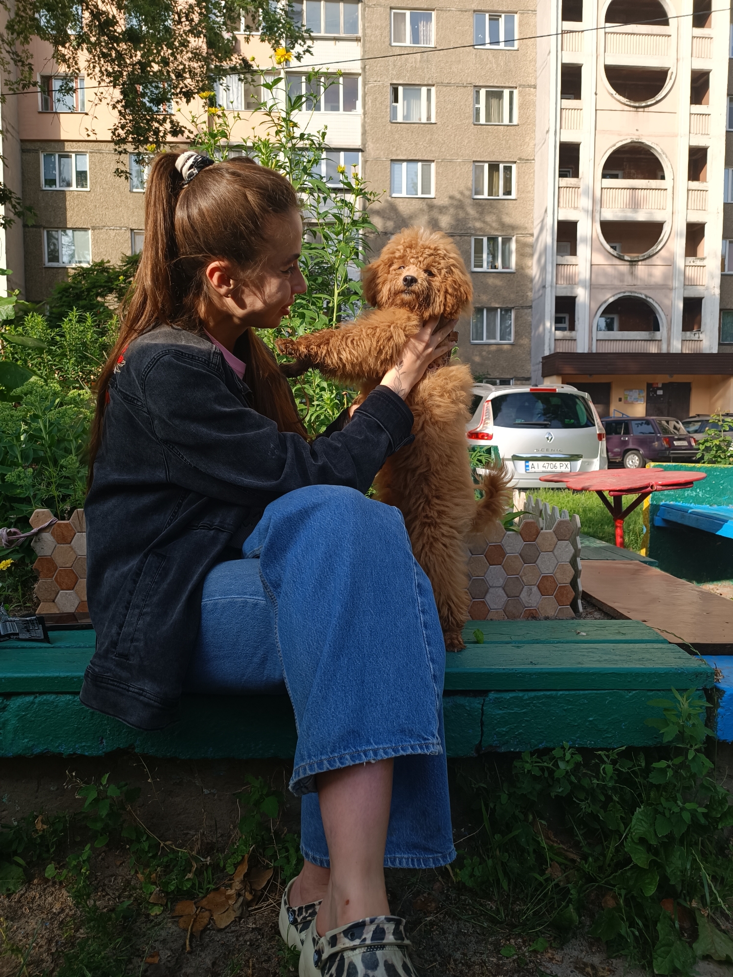 A young woman with long brown hair tied in a ponytail is sitting on a green wooden bench and holding a small brown dog. The woman is wearing a dark denim jacket over a pink shirt and blue jeans. The dog appears to be a poodle mix. They are in an outdoor setting with a building in the background, green foliage, and a sandpit. The building is a multi-story apartment complex with balconies and windows. A car is parked in front of the building. The lighting suggests it is daytime.