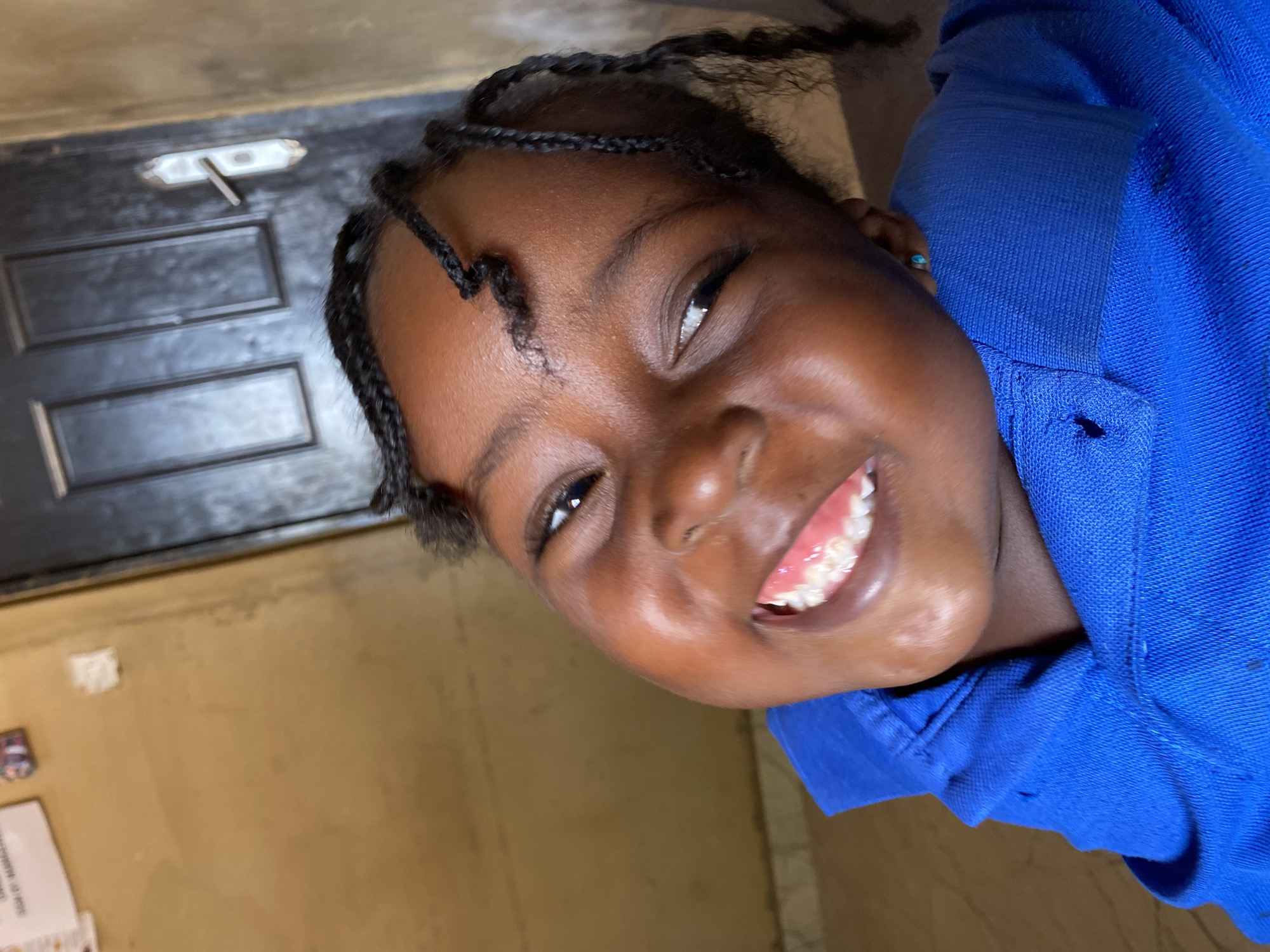 The media file is a close-up, diagonal portrait of a young Black girl, smiling broadly and looking directly at the viewer. Her face is bright with a wide, joyful smile revealing her teeth, and her eyes are open and engaging. Her dark skin has a natural sheen, and her hair is styled in numerous small, intricate braids that frame her face. She is wearing a vibrant royal blue top, which appears to be a polo shirt based on its textured fabric and visible collar. A small, light blue stud earring can be seen in her right ear.
The background suggests an indoor setting. Behind her head and upper body is a dark brown or black door, featuring two recessed panels and a simple, metallic-colored latch or handle mechanism at the top. The rest of the background consists of a plain, light brown or tan wall. On this wall, in the lower left portion of the image, there is a small, blurry piece of white paper or material with faint, indiscernible text. The overall atmosphere conveyed by the girl's expression is one of happiness and warmth. The lighting suggests an interior space, but no specific time of day or weather is evident.