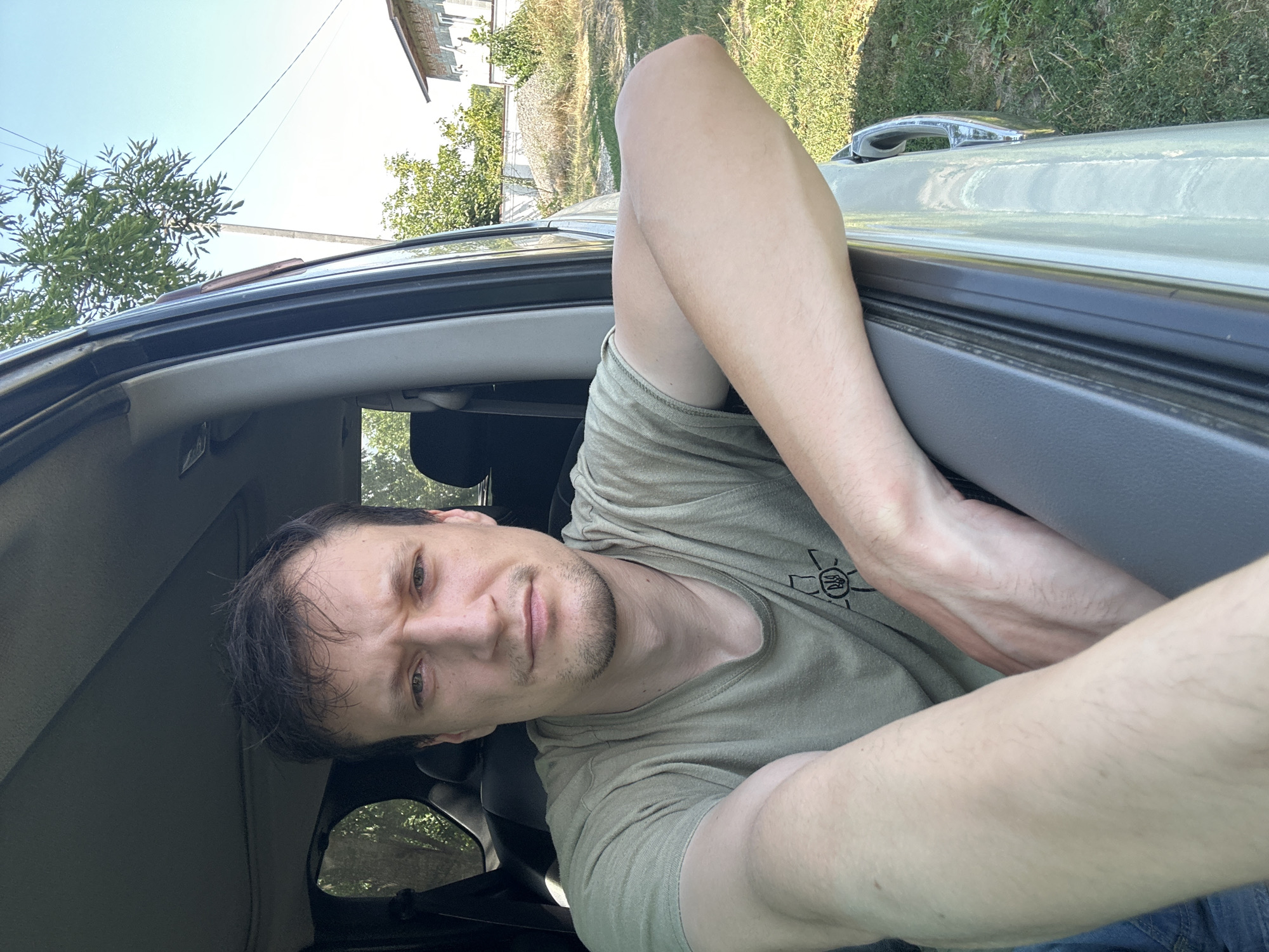 A man is pictured in the driver's seat of a vehicle, leaning out the open window. He is looking directly at the camera with a neutral expression. He is wearing a light olive-green t-shirt with a symbol on the left chest. His right arm is resting on the car door frame, and his left arm is bent across his chest. The background shows greenery, a house, and a clear blue sky, suggesting a rural or suburban outdoor setting. The specific location within Makiv, Ukraine, cannot be determined from the image alone.