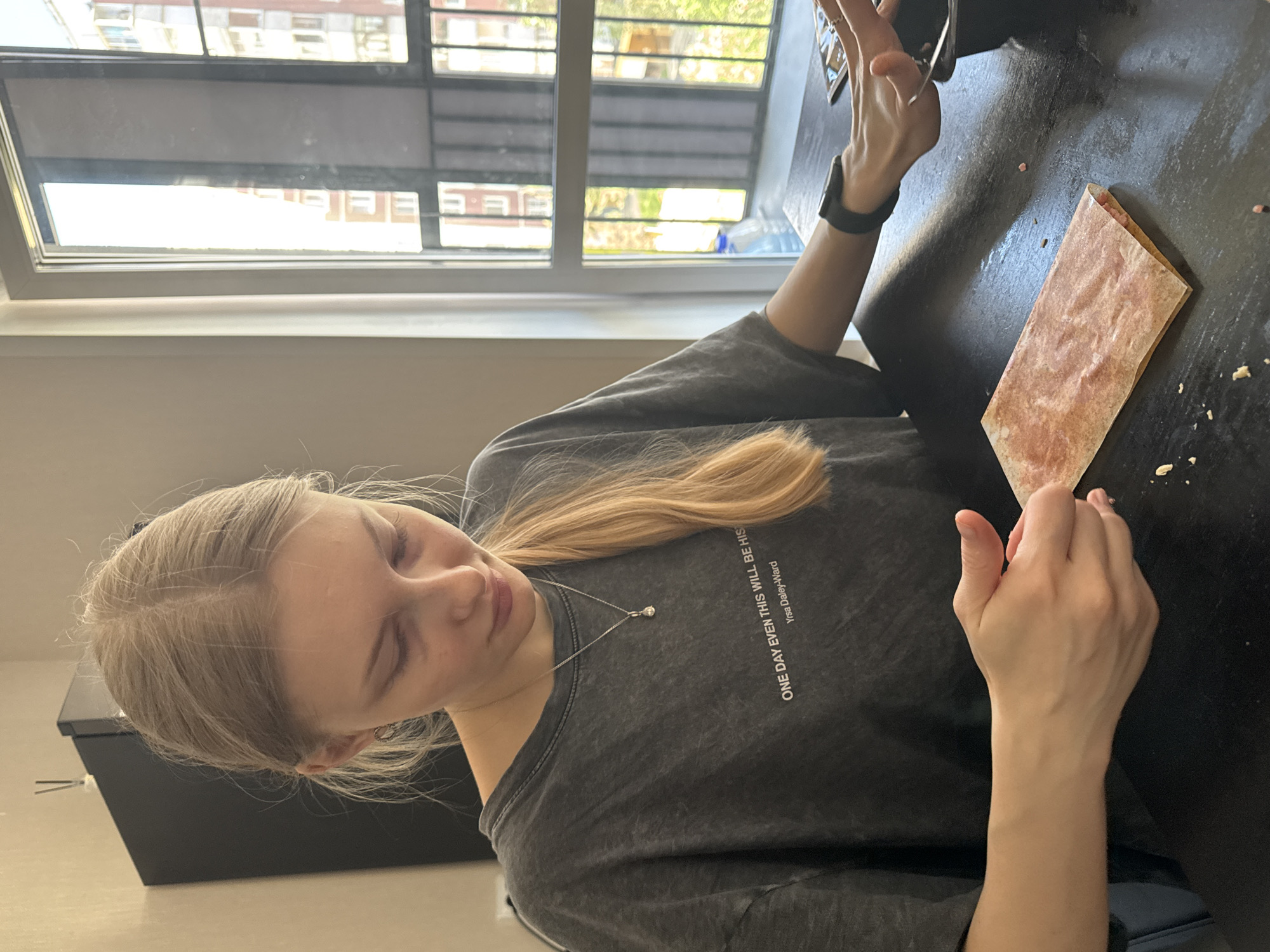 A young woman with blonde hair is seated at a dark-colored table in what appears to be an indoor setting. She is wearing a gray t-shirt with text on it, and a silver necklace with a pendant. She is holding a smartphone in her left hand and a folded piece of food or paper in her right hand. The table shows crumbs or small debris. A window is visible in the background, showing a building exterior. The location appears to be a city in Russia, and possibly Perm.