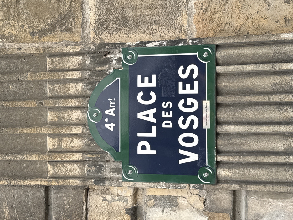 A Parisian street sign, primarily dark blue with white lettering and a green border, is vertically centered on an aged, light-colored stone wall. The sign identifies the location as "PLACE DES VOSGES". An arched section at the top of the sign indicates "4ᵉ Arrᵗ", signifying the 4th Arrondissement in Paris, France. A smaller, handwritten inscription, "Je pense toujours à toi, Rachel," is visible at the bottom edge of the main sign. The stone wall features prominent vertical fluted or reeded architectural elements, displaying signs of weathering, minor erosion, and discoloration consistent with historical construction. The sign is secured to the wall by four visible silver-colored fasteners, each with a spiral-like design.