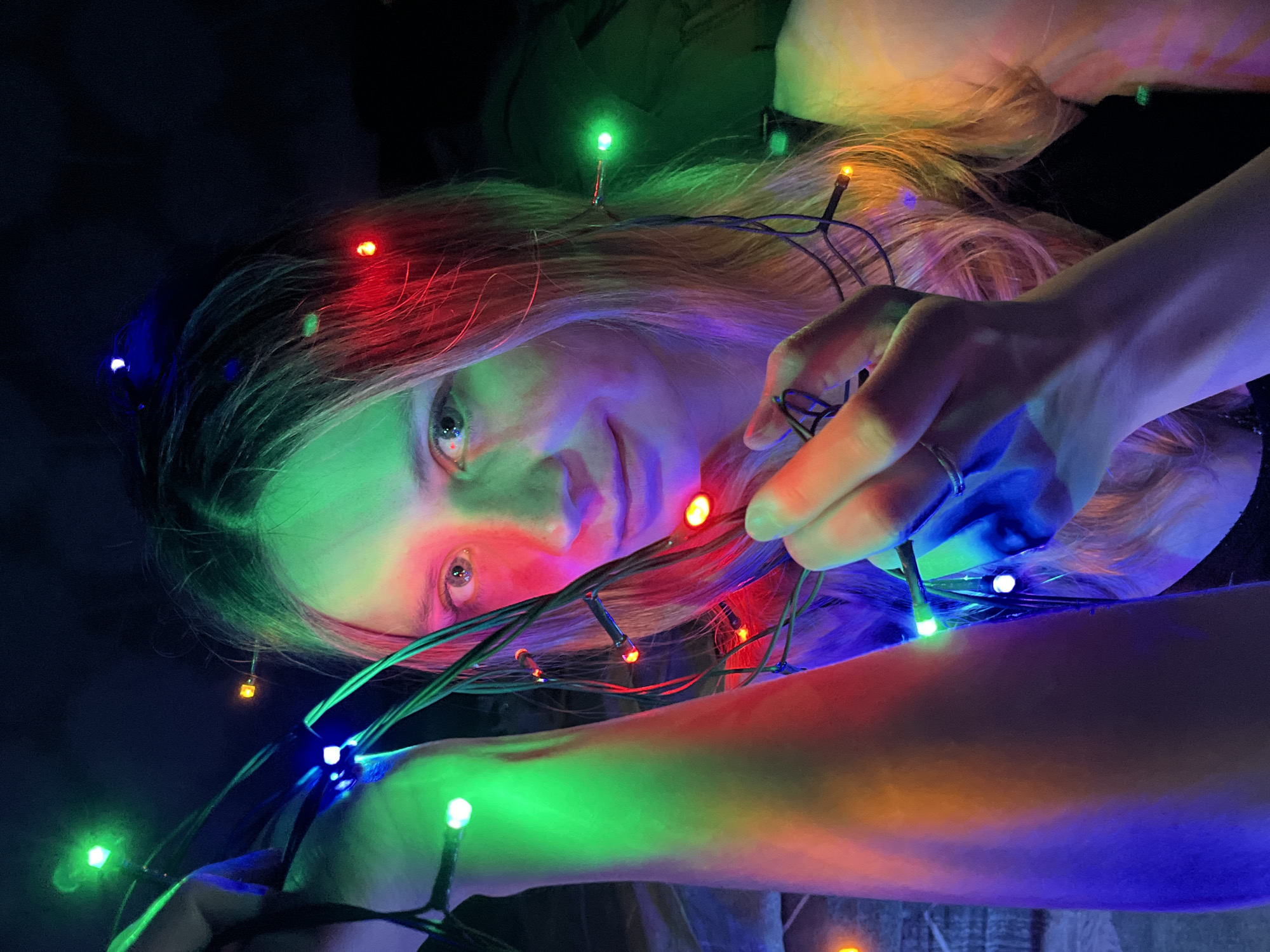 A young woman lies against a dark background, possibly indoors, holding a string of multicolored Christmas lights. The lights are draped around her head and arms, casting colorful reflections on her face and skin. The light colors include red, green, blue, and yellow. The woman has long, fair hair. She is wearing a ring on her finger. There is no information to confirm this image was taken in the city of Chaikovsky, Russia.