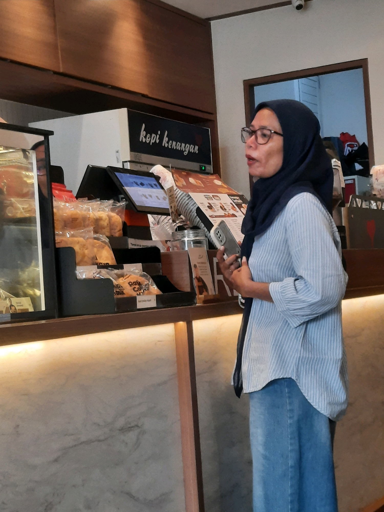 In East Jakarta, Indonesia, a woman wearing glasses and a dark blue hijab is standing near a display of packaged snacks. She is also wearing a light blue and white striped shirt and blue jeans. In her right hand, she is holding a smartphone. Behind her, there is a menu or display board with text in Indonesian, including the words "kopi kenangan". The background shows a counter with a marble-like pattern, and a wooden shelf with lighting above it. The scene appears to be inside a cafe or food establishment.