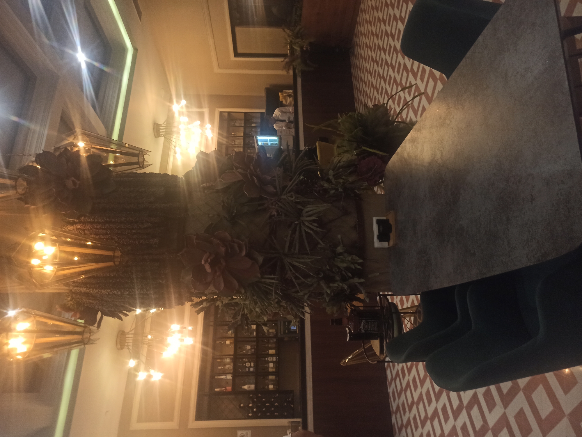 The image shows the interior of a restaurant or bar. In the foreground, a large, dark gray rectangular table with a textured surface is angled towards the right. Several dark teal chairs are visible to the left of the table, with their backs slightly curved. Behind the table, a large, decorative pillar covered in artificial foliage and large, dark red floral elements is prominent. Further back, a bar area is visible with shelves stocked with bottles of alcohol. A bartender in a white shirt and tie stands behind the bar. The floor has a distinctive geometric pattern of beige and red squares. In the background, several modern chandeliers with warm lighting are suspended from the ceiling. The overall lighting is warm and ambient.