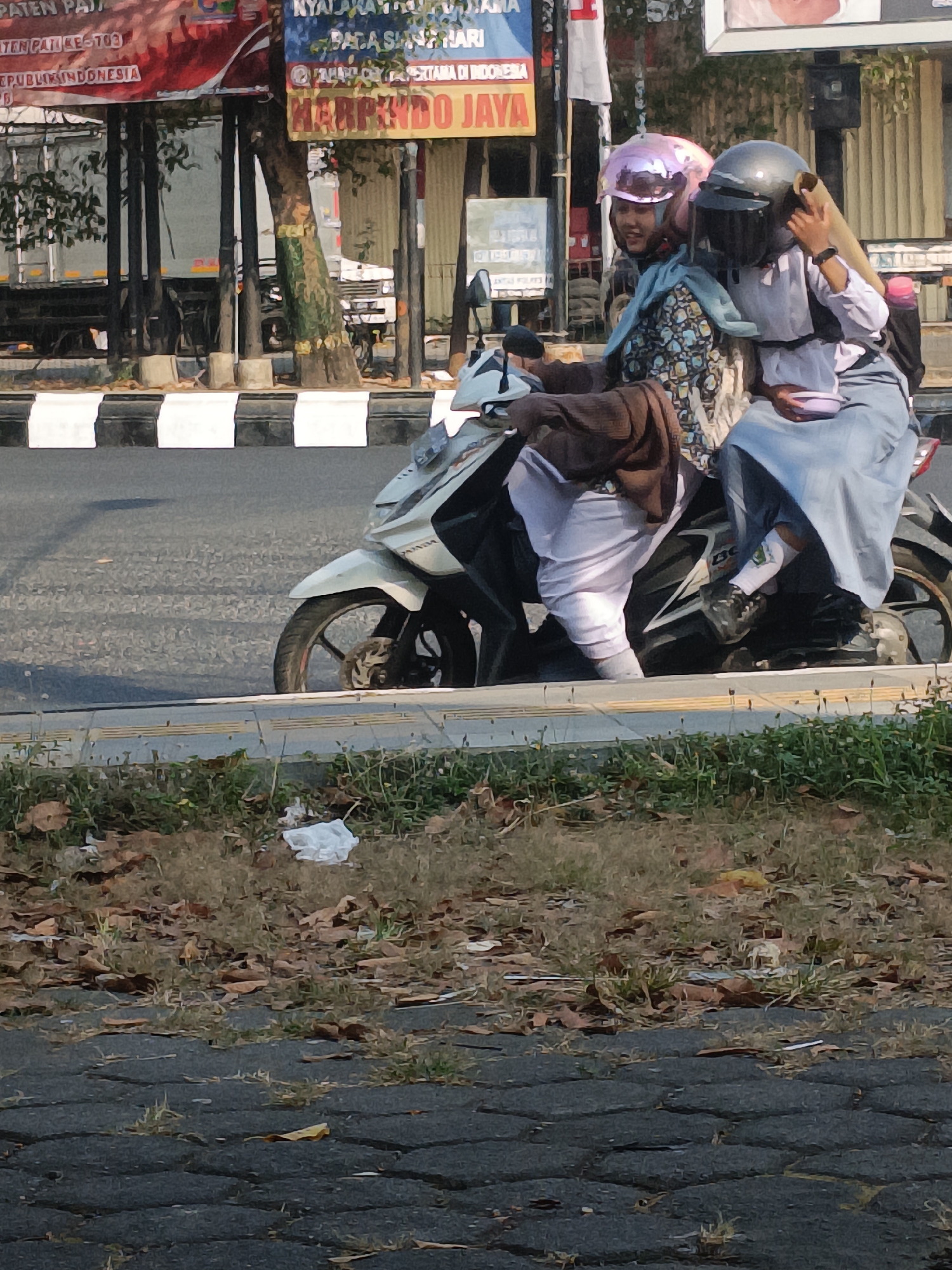 Three individuals, two female and one male, are riding on a white motorcycle. The motorcycle is in motion on a paved road. The two females are seated in the back and the male is in the front, driving. All three are wearing helmets. The female in the very back is wearing a pink helmet, the female in the middle is wearing a dark gray helmet, and the male in front is wearing a dark gray helmet. They are all dressed in school uniforms. The image is taken from the side, showing the motorcycle in profile. In the background, there are several billboards, trees, and a truck. The scene appears to be in a busy urban area.