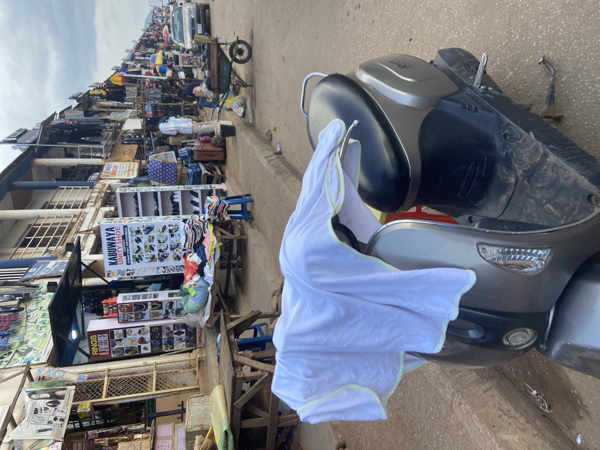 The image depicts a street scene in Bukuru, Nigeria, likely a market area. In the foreground, a grey scooter is parked, covered with a white cloth that has a light green trim. The scooter is positioned on a dirt road. Behind the scooter, the street is lined with shops and stalls selling various goods, including clothing, shoes, and jewelry. Several people are visible in the background, some attending to their businesses and others walking. A wheelbarrow filled with goods, and a car are also present on the street. The environment appears to be an outdoor market with a mix of commercial activity and everyday life. The buildings have a rustic appearance, and the overall atmosphere suggests a bustling, informal marketplace.