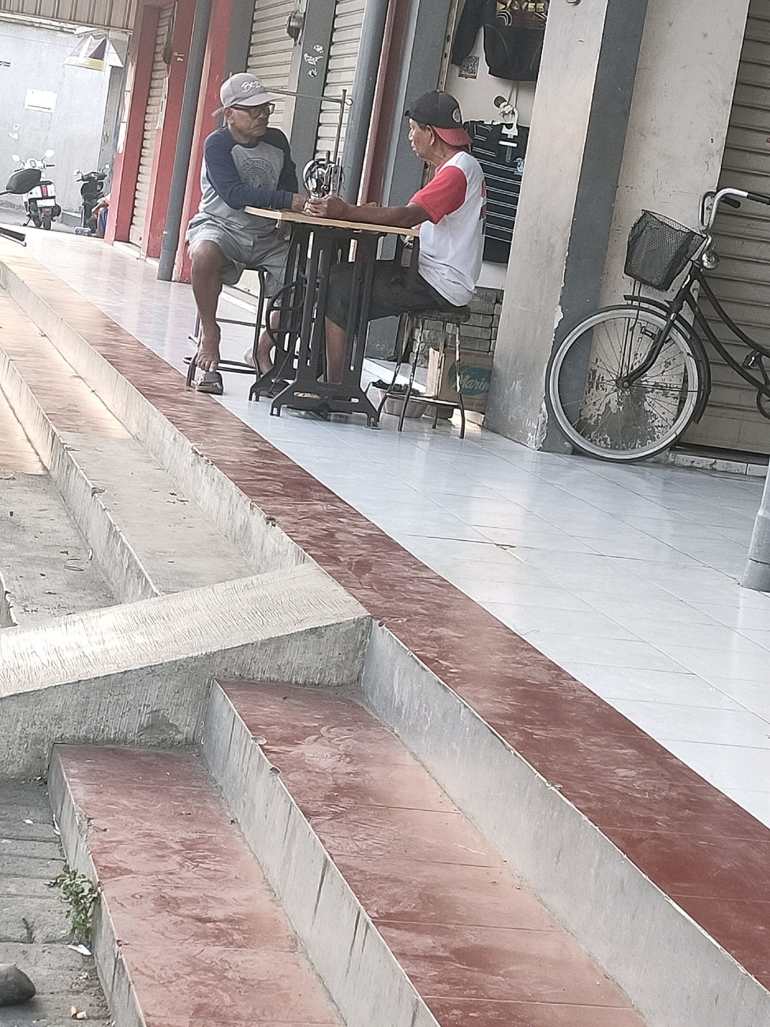 Two men are in front of shops. One man on the left is older, wearing a grey long-sleeved shirt, grey shorts, glasses, and a grey baseball cap. He is sitting on a stool, with his left foot on a sandal, and his right foot bare. He appears to be operating a sewing machine. The other man on the right is younger, wearing a red and white t-shirt and a black baseball cap, and is seated at the same table as the older man, looking towards him. A sewing machine is on a table between them. In the background, there are scooters and a bicycle with a basket. The scene appears to be outdoors on a tiled walkway with concrete steps.