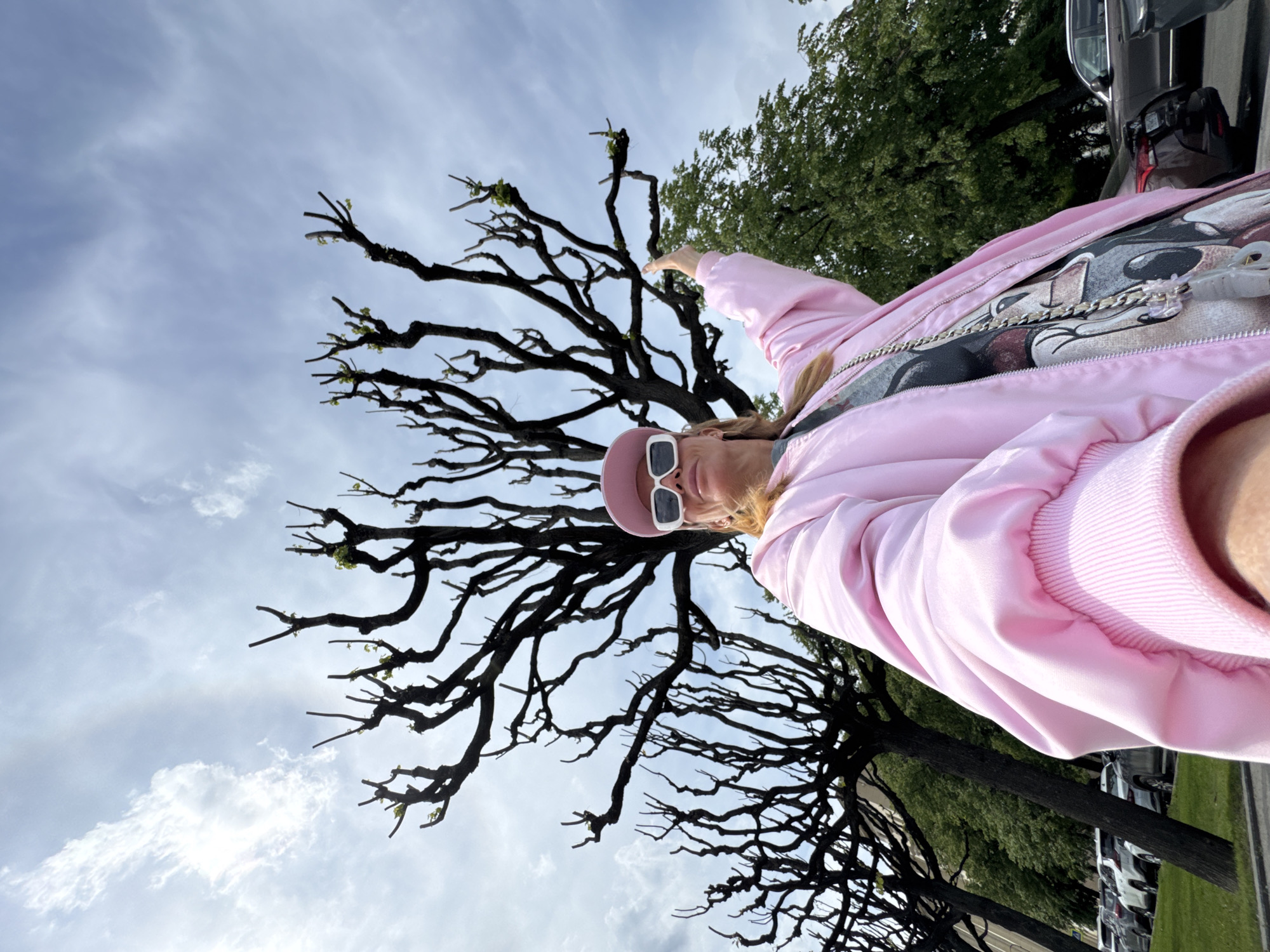 The image depicts a person taking a selfie outdoors in Saint Petersburg, Russia, under a partially cloudy sky. The person, appearing to be female, is positioned in the foreground, wearing a bright pink bomber jacket, white rectangular sunglasses, and a matching pink baseball cap. Beneath the jacket, a dark t-shirt with a graphic print of a cartoon character, possibly Mickey Mouse, is visible, accented by a silver chain worn around the neck. The person has blonde hair and is looking directly at the camera.
The background is dominated by large, deciduous trees with intricate, dark, and mostly bare branches, from which small green buds are just beginning to emerge, indicating early spring. Further in the background, on the right side of the frame, there are trees with full, vibrant green foliage. To the far right, a segment of a parking lot with several cars and a strip of green grass is visible. The scene is brightly lit by daylight. There is no visible text in the image.