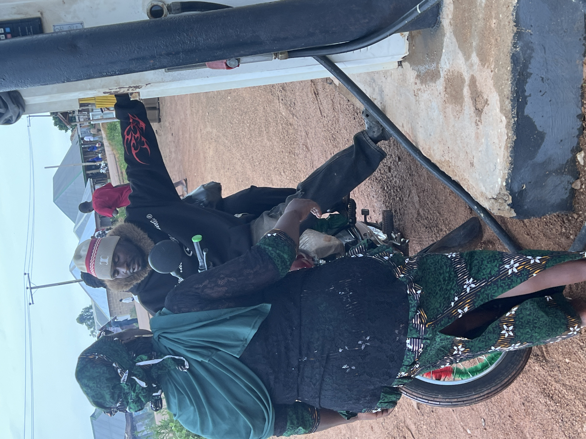 In Durumi, Nigeria, at a gas station, a woman wearing a green headscarf and a black lacy dress with a green patterned skirt is standing next to a motorcycle. A man, wearing a beanie with red stripes and a black jacket with red flame decals, is sitting on the motorcycle and interacting with the woman. The man appears to be fueling the motorcycle from a pump on the right side of the image. The ground is made of dirt and gravel, and in the background, there are several buildings and people. The sky is overcast.