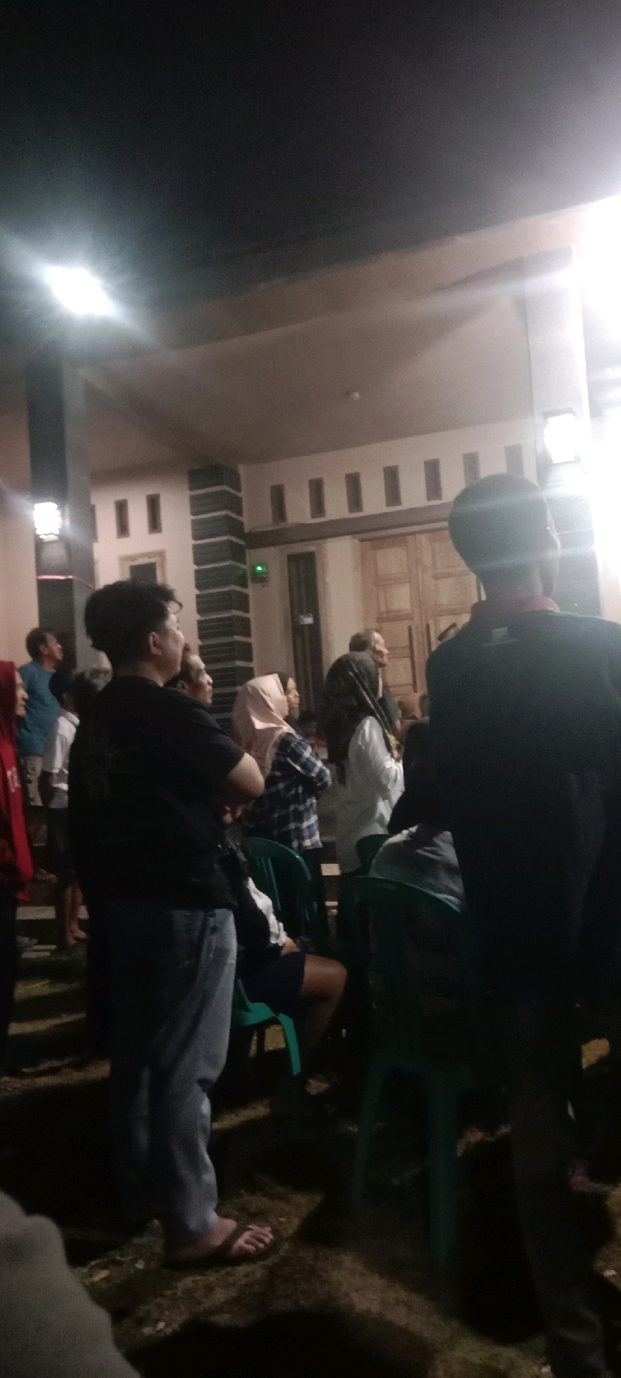 The image depicts an outdoor gathering at night, potentially in the city of Kras, Indonesia. A group of people are standing and sitting, illuminated by artificial lights. In the foreground, a person in a black t-shirt and jeans is visible, facing away from the camera. To their left, another individual in a blue shirt is seen looking upwards. Several other individuals are dispersed throughout the scene, some wearing headscarves. Green chairs are placed on the ground, with some people seated in them. The background shows a building with architectural features suggestive of Indonesian design, including wooden doors and decorative elements on the facade. The ground appears to be a mix of grass and dirt. The overall atmosphere suggests a casual social event.