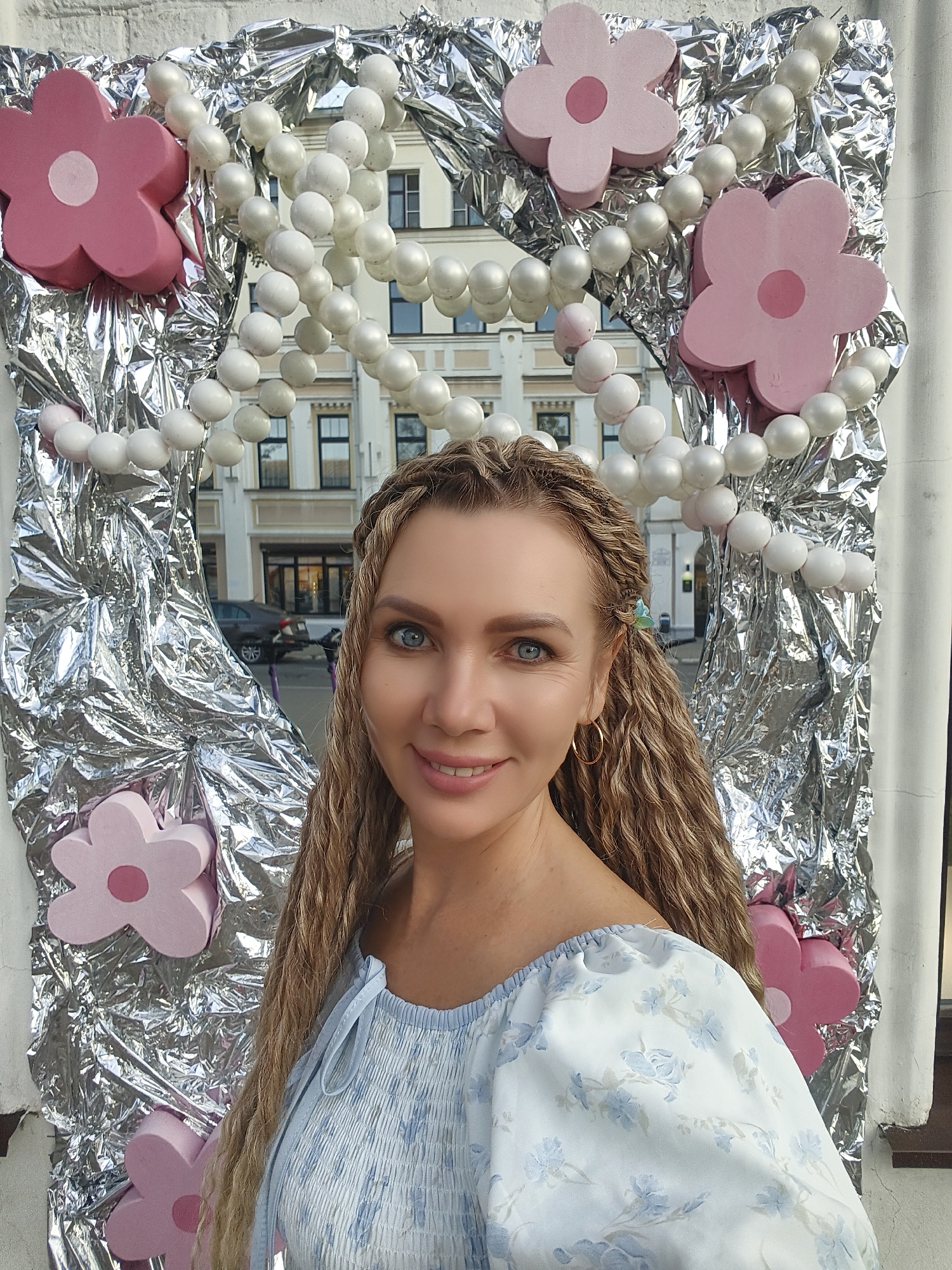 A medium shot shows a woman with braided blonde hair and blue eyes smiling and looking at the camera. She is wearing a blue and white floral print off-the-shoulder top. The background is a silver foil-covered structure decorated with white bead strands and pink flowers. Buildings are visible through a gap in the decoration. The woman is positioned in front of this backdrop, which appears to be an outdoor urban setting.