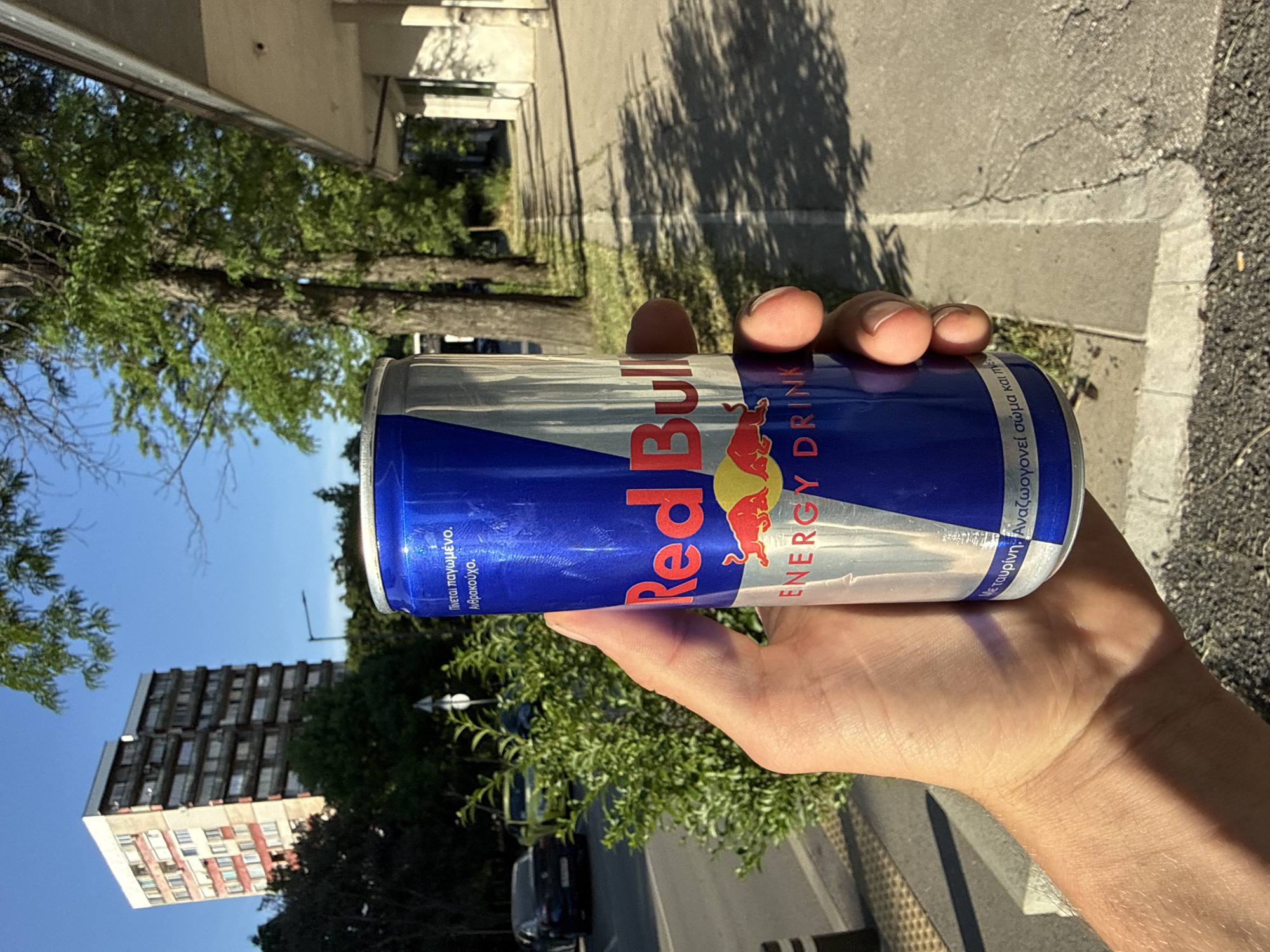 A person's hand holds a Red Bull energy drink can outdoors in the daytime. The can is predominantly silver with blue and red branding. The background shows a tall, multi-story building with balconies, trees with green leaves, a sidewalk, and a street with parked cars. The lighting suggests a sunny day.