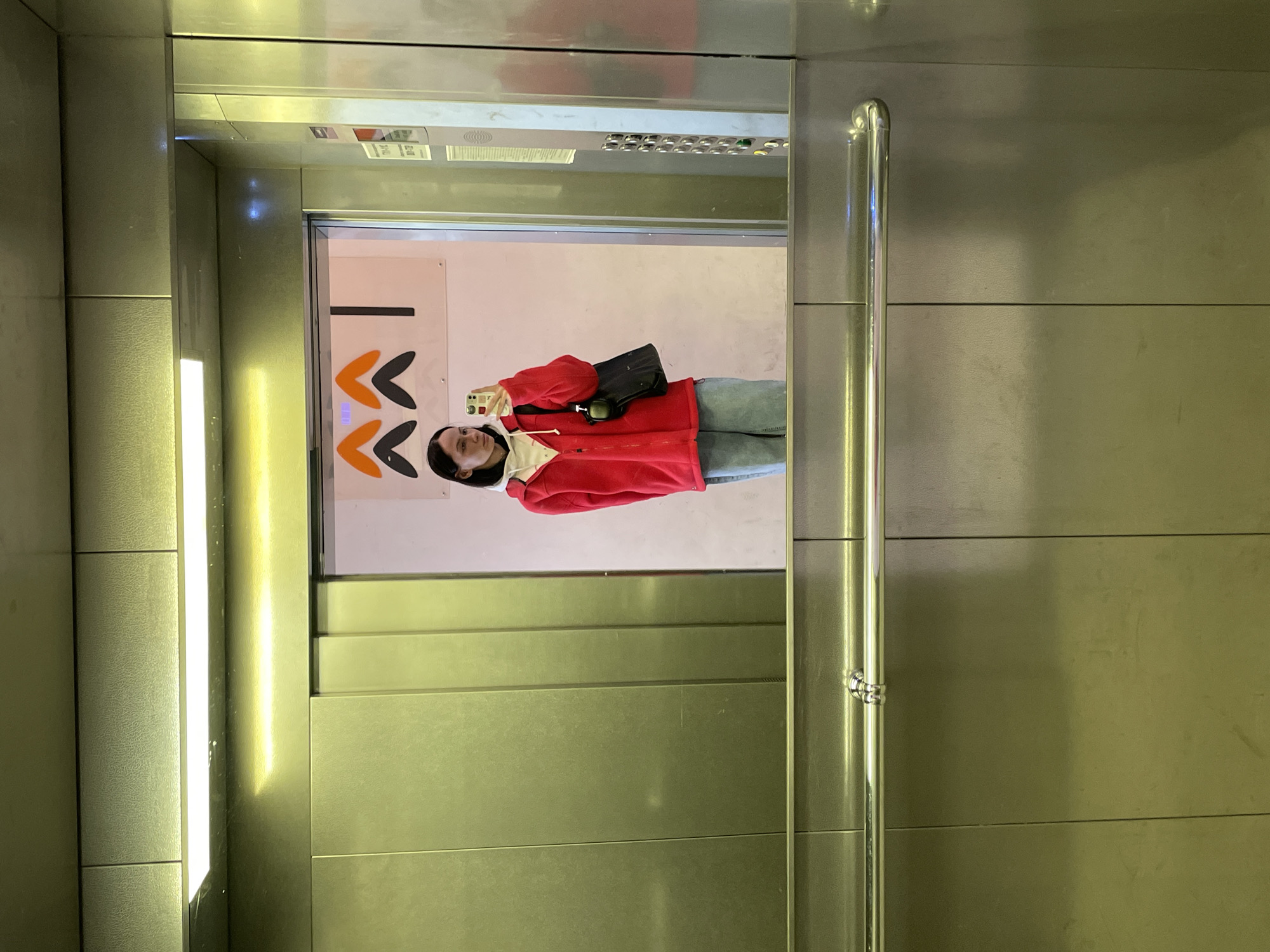 The scene depicts a mirror selfie taken inside an elevator. The location is an elevator within a building affiliated with Axion Holding (Аксион Холдинг) in Izhevsk, Russia, identifiable by the distinctive orange and black upward-pointing chevron arrow logo and a vertical black line visible on a sign reflected through the partially open elevator doors.
A single female subject is present, centered in the reflection. She has dark, straight hair and a neutral expression while looking at the smartphone in her right hand, which has a white case and is positioned to capture the image. She is dressed in a white hooded sweatshirt layered under an unzipped red fleece jacket and light blue jeans. A black shoulder bag hangs from her left shoulder, and a small dark object is attached to her red jacket near her waist.
The elevator interior features brushed metallic panels on the walls and a horizontal, polished metal handrail running across the lower portion. Overhead, a rectangular fluorescent light fixture illuminates the space. To the right, a silver-toned control panel with a vertical array of circular buttons, including a green button, is visible. The reflection through the elevator doors shows a light-colored wall featuring the aforementioned corporate branding.