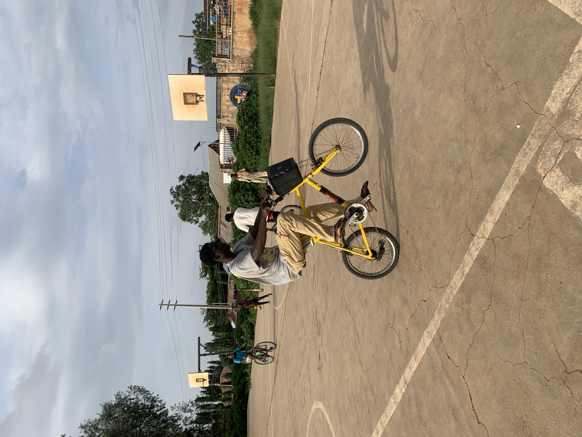 A young Black male is performing a wheelie on a yellow bicycle on a cracked concrete basketball court. He is wearing a grey t-shirt and tan pants. The bicycle has a black basket attached to the handlebars. In the background, another Black male is riding a bicycle, and a young Black child is running. Further in the background, there are basketball hoops, buildings, and trees. The sky is cloudy. The scene appears to be located in Jos, Nigeria, as indicated by the presence of a "Fellowship of FCA" sign on a building behind the basketball hoop.
