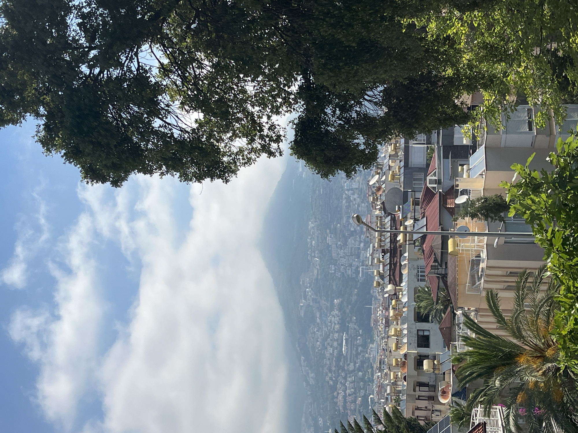 The image captures a vibrant, sunlit daytime scene of a city nestled against a mountainous backdrop, framed by lush green trees. The overall impression is bright and clear, with a few white clouds scattered across a blue sky.
In the immediate foreground, dense green foliage from trees frames the top and bottom sections of the view, suggesting the vantage point is from an elevated or tree-lined area. Sunlight filters through the leaves, highlighting some of the vibrant green.
The midground reveals a densely packed urban landscape, with numerous multi-story buildings stretching across a hillside. These structures feature varying architectural styles, many with balconies and reddish-brown tiled roofs. A prominent detail is the abundance of satellite dishes and what appear to be solar water heaters on almost every rooftop, reflecting the sunlight. A tall streetlight pole is also visible within the city sprawl. Scattered among the buildings are various green trees, including distinct palm trees with their characteristic fronds, adding to the verdant feel of the city.
In the background, a large mountain range dominates the horizon, its peaks partially obscured by a light atmospheric haze or low-hanging clouds, creating a sense of depth and distance. The city extends far into the distance, climbing the slopes of these mountains.
The sky above is a clear blue, with scattered white cumulus clouds, particularly more prominent on the left side, indicating fair weather. There is no visible text or explicit activity taking place; it's a static landscape view. Given the context, this is a scenic view of Alanya, Turkey, on a bright, sunny day.