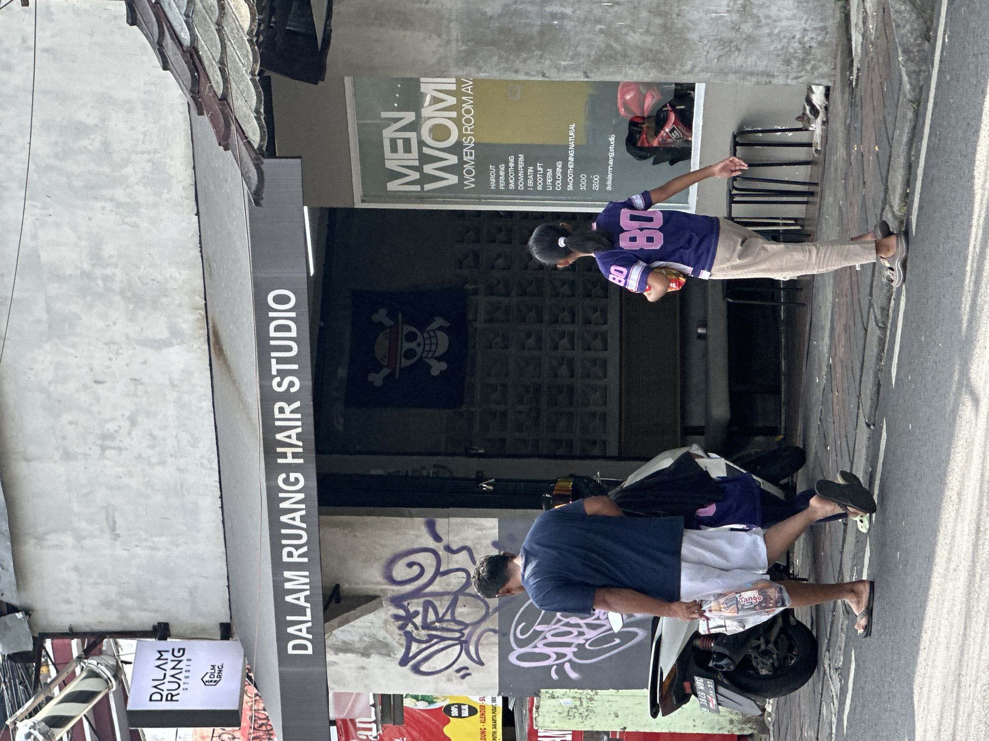 The scene shows the exterior of "Dalam Ruang Hair Studio" located in Central Jakarta, Indonesia. The storefront has a grey awning with black lettering. A sign displays the text "MEN... WOMEN'S ROOM AV" and lists services like haircuts, perming, and coloring. A black flag with a skull and crossbones is hanging in the background.
Two people are visible: a woman in a purple and white jersey is standing near the entrance, and a man in a blue shirt and white shorts is standing by a parked scooter. The man is holding a bag of Tango wafers. The scooter has a license plate that reads "B 3806 WBG". Graffiti is visible on the wall. The surroundings suggest a typical urban street setting.