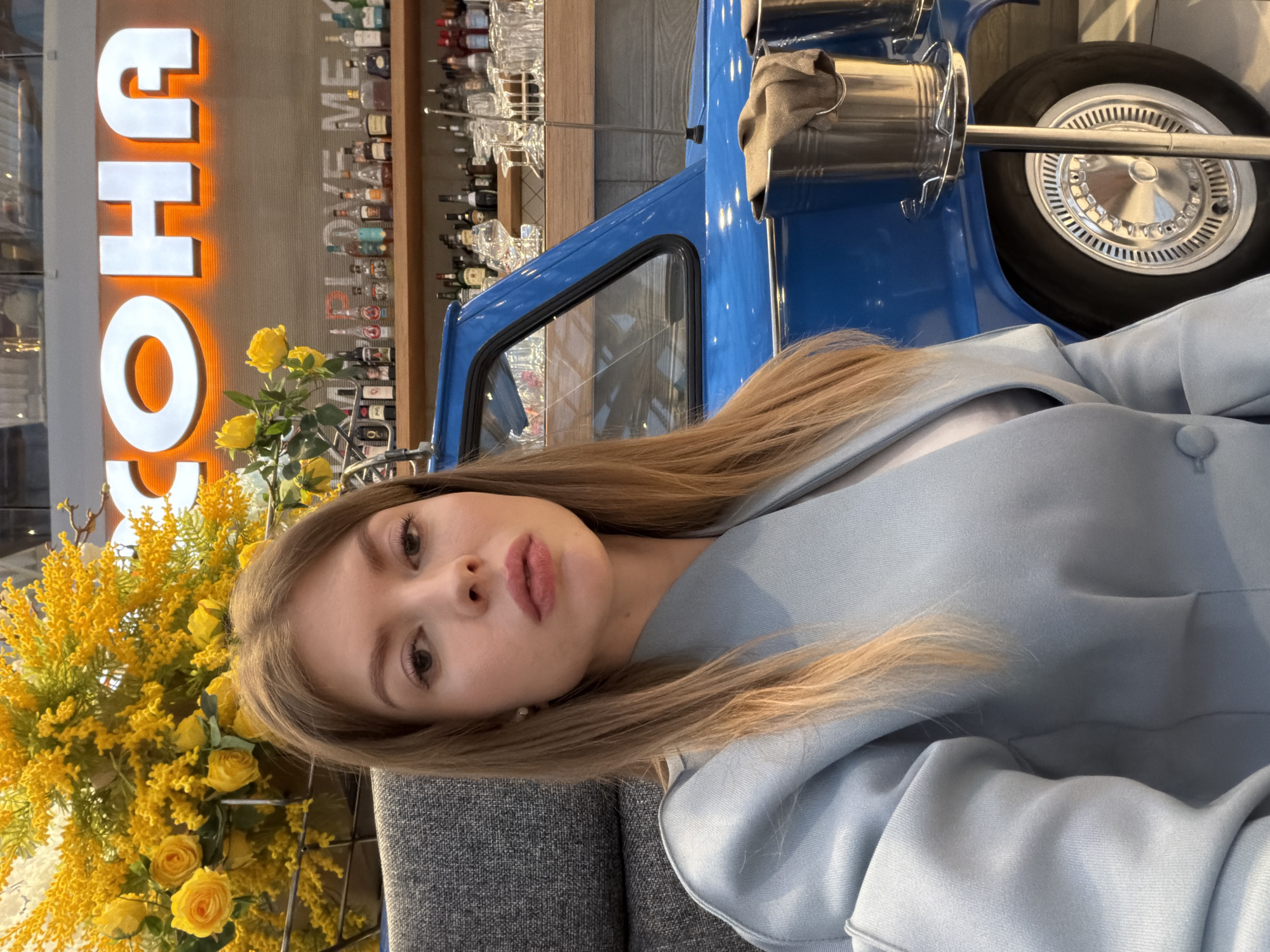 A young woman with long, light brown hair, dressed in a light blue blazer or jacket, is seated on a grey upholstered surface, looking directly at the camera with a neutral expression. The image is taken indoors, likely within a modern restaurant or bar in Khimki, Russia.
To her left, a large, vibrant arrangement of yellow flowers, including roses and other small blossoms, is prominently displayed. In the background, large, illuminated white letters spelling "OHS" (part of a larger sign, possibly "CHOOCHO") are visible. Below these letters, a fully stocked bar area extends, featuring numerous bottles of various spirits and glassware arranged on shelves. Faint text reading "LOVE ME" can be seen below the bar bottles.
On the right side of the frame, a significant portion of a bright blue vintage-style vehicle, possibly a decorative piece or a functional bar setup, is visible. It features a chrome wheel with a detailed hubcap and a small metal bucket attached to its side. The overall scene suggests a stylish and well-lit commercial space.
