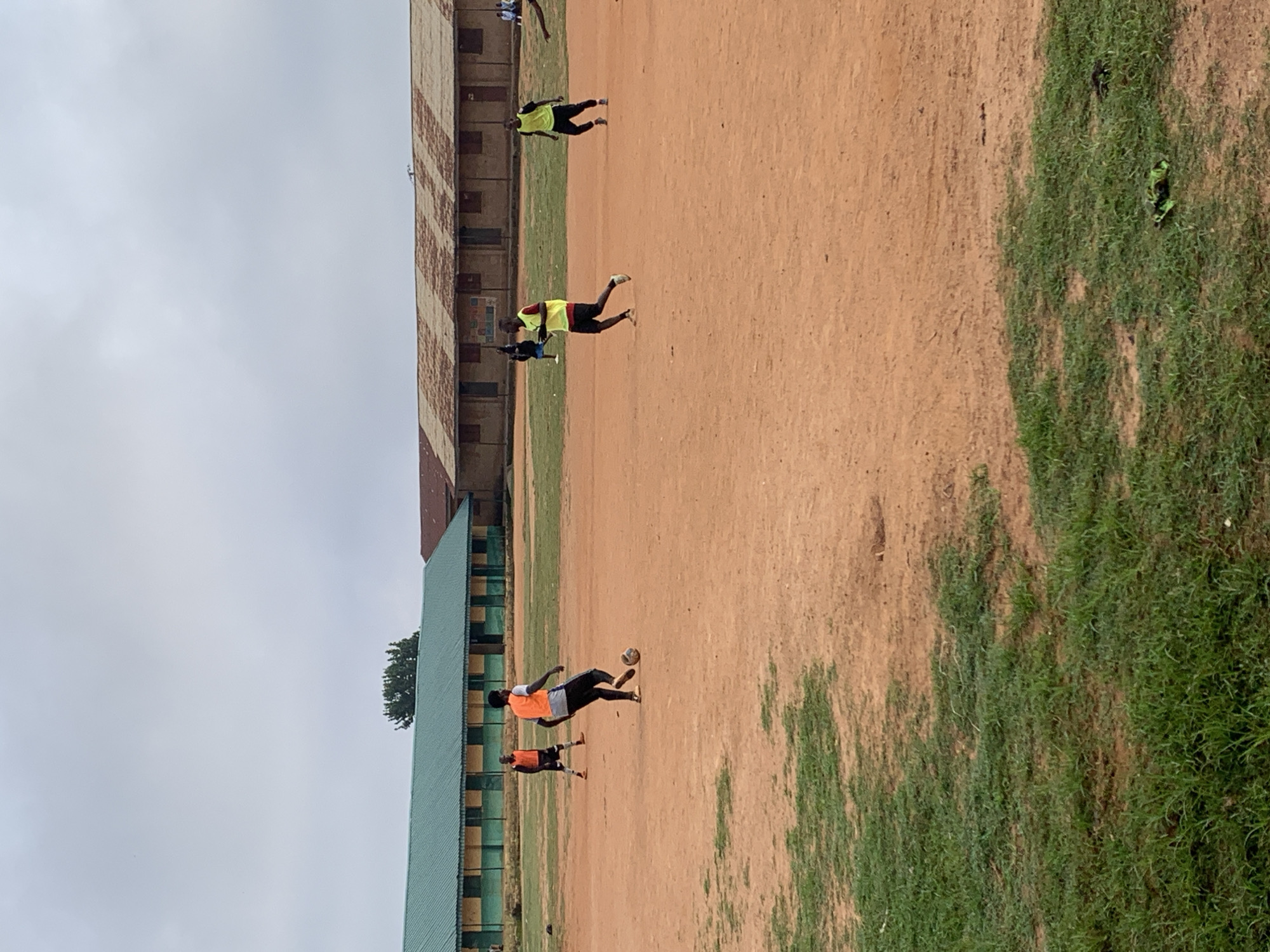 The image depicts an outdoor football field in the city of Bukuru, Nigeria. The field is predominantly made of red dirt with patches of green grass around the edges. In the background, there are two long buildings with green and brown corrugated roofs respectively. The green-roofed building appears to be a school or administrative block, with multiple windows and an open-air walkway. The brown-roofed building is older and shows signs of wear.
Several individuals are present on the field, engaged in a football game. One person in an orange vest is kicking a soccer ball. Another person in an orange vest is standing further away on the field. Two individuals wearing yellow vests are also visible, one running and the other in motion. The sky is overcast, suggesting a cloudy day. The overall scene captures a moment of casual sporting activity in a semi-urban environment.