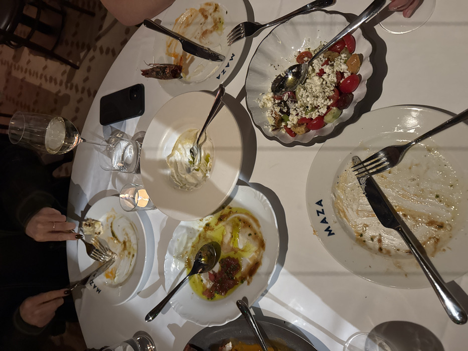 The image displays an overhead view of a white-clothed table within a dimly lit restaurant named MAZA. Multiple white plates, several bearing the "MAZA" logo, are present, predominantly showing remnants of consumed food. In the upper left, a person's hands, one with a wedding band, are actively cutting food on a plate using a knife and fork. Other plates contain residue such as olive oil and tomato, a creamy white dip, a shrimp head, and a salad composed of cherry tomatoes, cucumber, croutons, and crumbled feta cheese. Utensils, including knives, forks, and spoons, are scattered among the plates. A wine glass with white wine, two water glasses, and a black smartphone are also on the table. A single lit tea light candle in a glass holder provides central illumination. The background shows blurred elements of a restaurant interior, including patterned flooring and dark chairs.