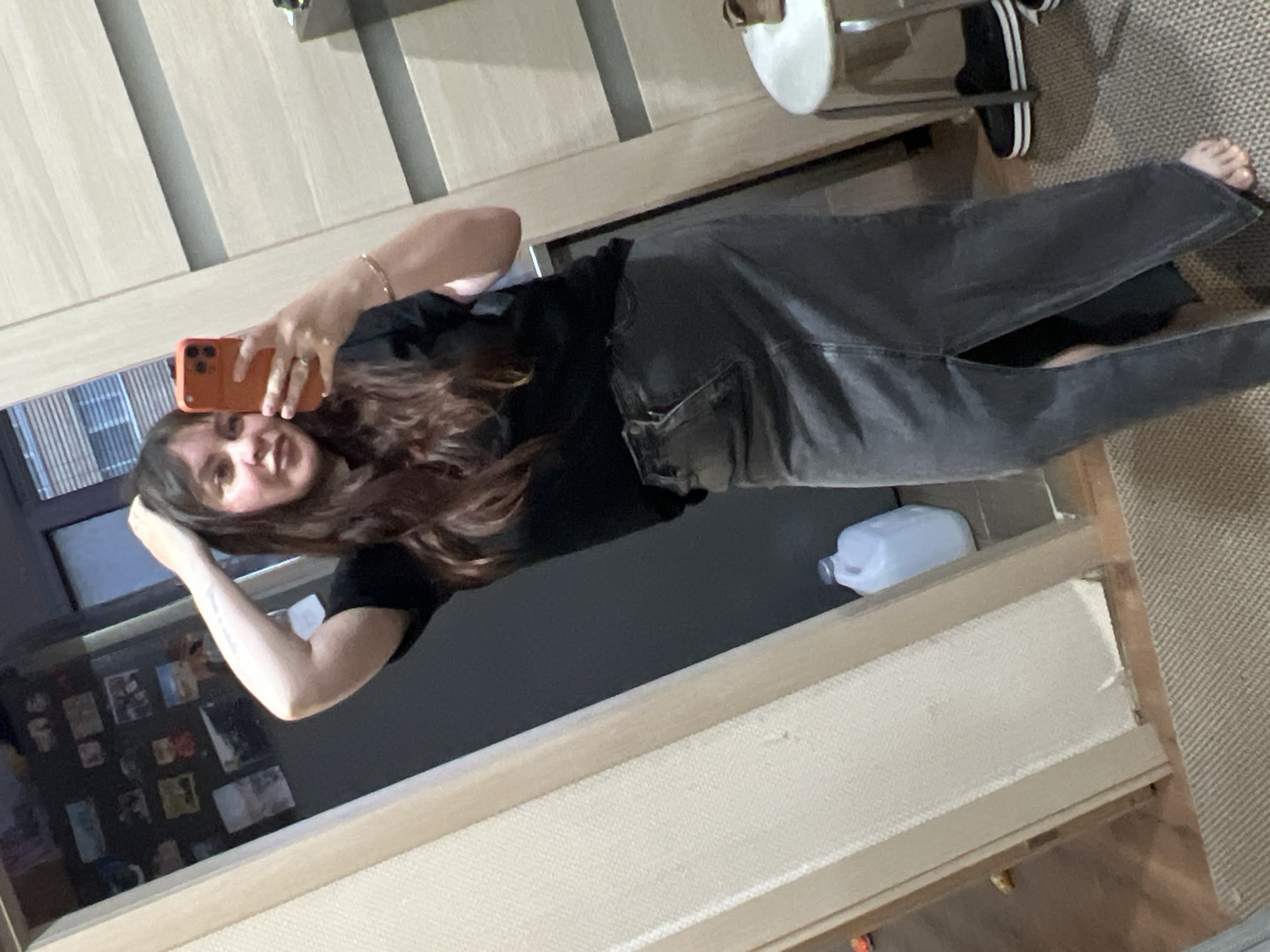 The media file shows a young woman taking a mirror selfie in what appears to be a bedroom or dressing area in Noviy, Russia. She is wearing a black t-shirt and dark grey jeans. She is holding an orange smartphone in her right hand, and her left hand is resting on her head. Her hair is long and brown, and she has a slight smile on her face, suggesting a casual and relaxed mood.
In the background, the room is furnished with wooden cabinets, indicating a domestic setting. The floor appears to be carpeted in a neutral tone. There are several framed pictures visible on a shelf or surface, suggesting personal adornment of the space. A white plastic jug is also visible in the background, and a pair of black and white sneakers are on the floor. The lighting suggests it is daytime, but the specific time is not discernible. The overall impression is of a personal moment captured for social media or personal memory.