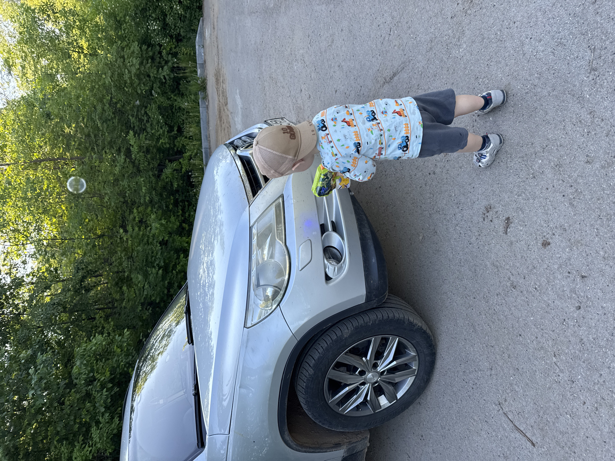 The image depicts an outdoor scene in Vereshchagino, Russia. A silver Volkswagen Tiguan SUV is parked on light grey asphalt pavement. The front driver's side of the vehicle, including the headlight and a multi-spoke alloy wheel, is prominently featured. A small child, viewed from the rear, stands facing the front bumper of the SUV. The child is wearing a light blue long-sleeved shirt patterned with cartoon vehicles (trucks, tractors, horses, trees), dark grey shorts, and grey and white athletic shoes. A light brown mesh baseball cap, displaying "10 GOS" text, is worn by the child. The child holds a green and yellow toy bubble gun, aiming it towards the vehicle's front bumper. A single, translucent bubble is suspended in the air in the upper left portion of the frame, above the SUV. The background consists of dense green deciduous trees and foliage, with a concrete curb separating the paved area from the treeline.