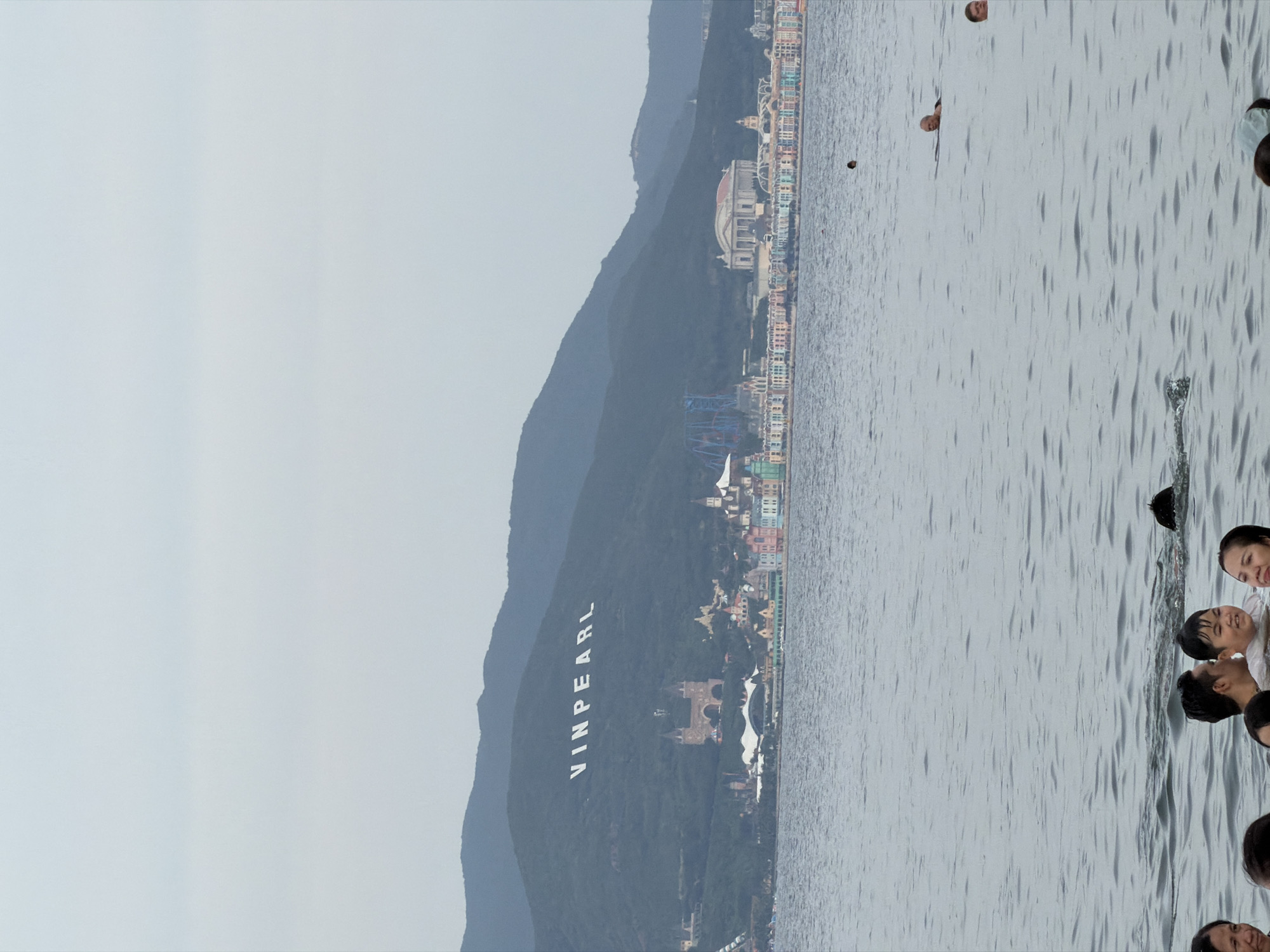 The image captures a wide shot of the ocean in front of a coastal city in Nha Trang Ward, Vietnam. In the background, a large sign spelling "VINPEARL" is visible on a mountainside, indicating the location of Vinpearl Land, a popular amusement park and resort complex. The city skyline features a collection of colorful buildings, including what appears to be a large classical-style structure and a roller coaster. The water in the foreground is populated by several individuals swimming and wading. A group of people are clustered in the lower part of the frame, appearing to be in shallow water. Further out, there are individuals swimming. The sky is overcast, casting a diffused light over the scene.