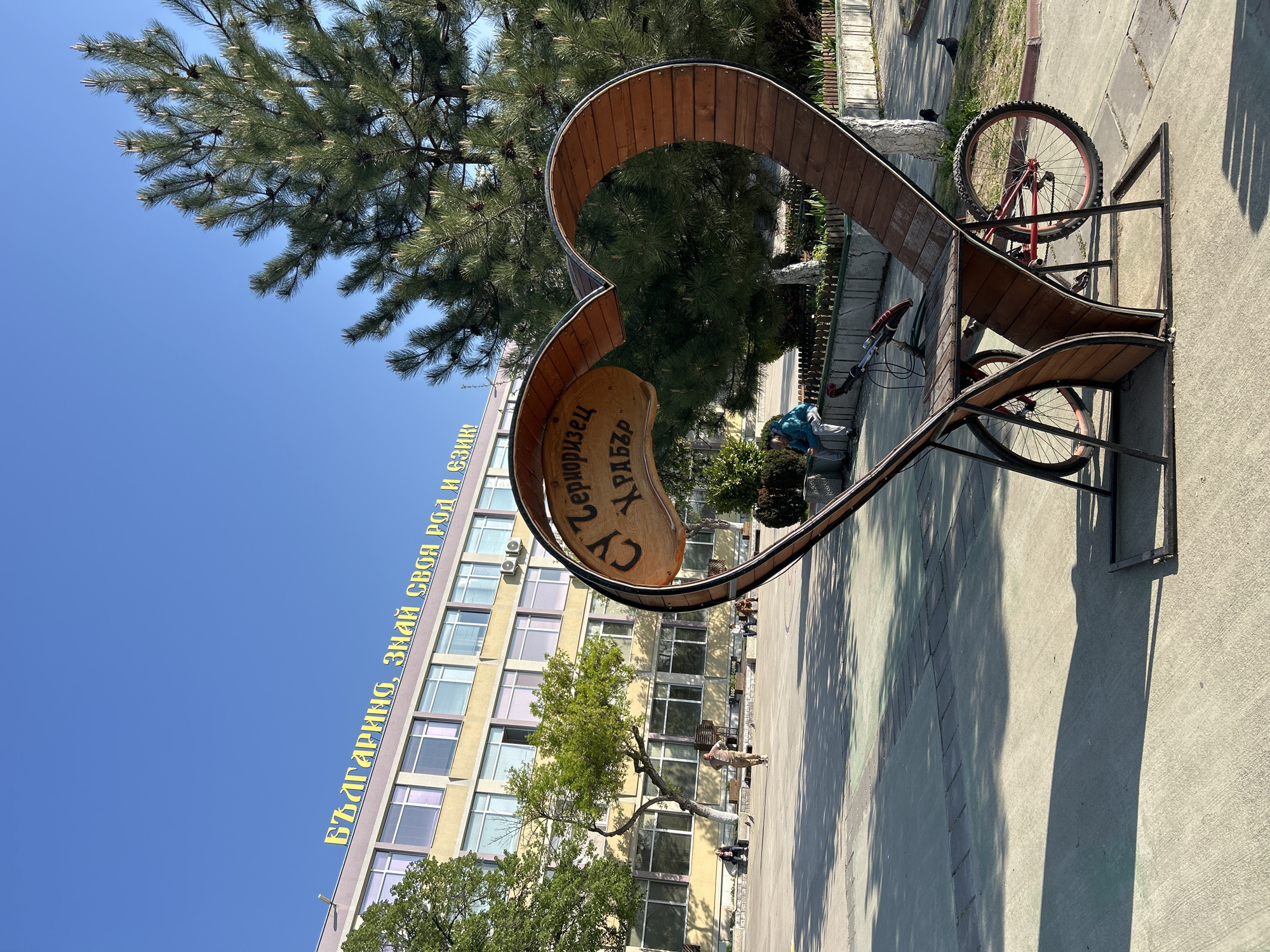 The image shows a heart-shaped sculpture in Plovdiv, Bulgaria. The sculpture is made of wood and metal and has a plaque inside it with text in Cyrillic script. The background includes a multi-story building with yellow and brown exterior, and a blue sky. There are people and trees visible in the background. A bicycle is parked on the sculpture. The scene is bathed in sunlight, casting shadows on the ground.