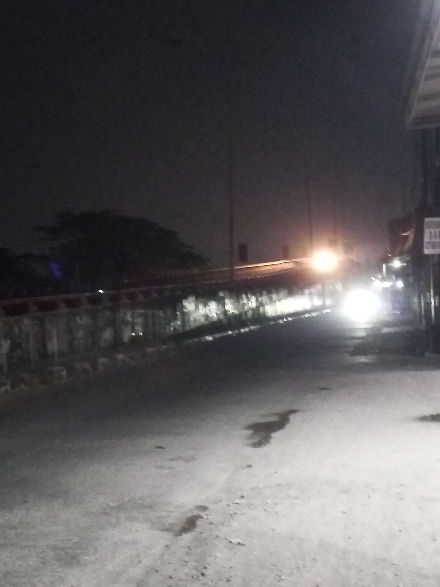 This image depicts a nighttime scene on a street in Gebang, Indonesia. The foreground shows a paved road with patches of water or wetness, and some debris scattered on the surface. In the mid-ground, a dark silhouette of a tree or dense foliage is visible against the sky, with a reflective barrier or railing running horizontally in front of it. Beyond this, a bright, blurred light source, possibly a vehicle's headlights or streetlights, illuminates the road ahead. To the right, a building with an awning or roof structure is partially visible, along with a vertical pole that may support a sign. The overall atmosphere is dark and somewhat grainy, suggesting low-light photography.