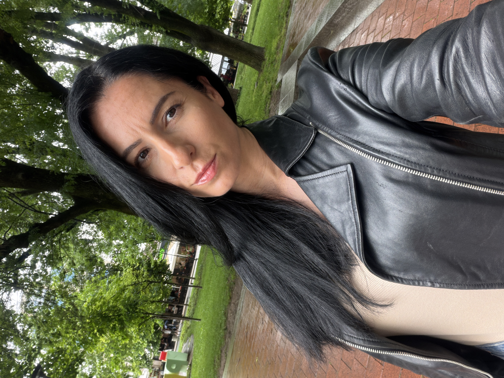 This selfie captures a woman outdoors in what appears to be a park setting in Pleven, Bulgaria. The subject is a woman with long, straight dark hair, dark eyebrows, and brown eyes, looking directly at the camera with a neutral to slightly serious expression and glossy lips. She is dressed in a black leather jacket with prominent silver zippers and distinctive ribbed detailing on the sleeves, worn over a light beige or tan top.
The background features lush green foliage from large trees, indicating a natural, possibly urban park environment during a warm season like spring or summer. The ground in the foreground and to the right is a damp, reddish-brown paved surface, suggesting recent rainfall. In the middle distance, behind the trees, parts of a building with windows are visible, possibly a cafe or restaurant, along with some indistinct colorful objects. The lighting is bright and diffused, consistent with an overcast daytime. No clearly legible text is visible in the image.