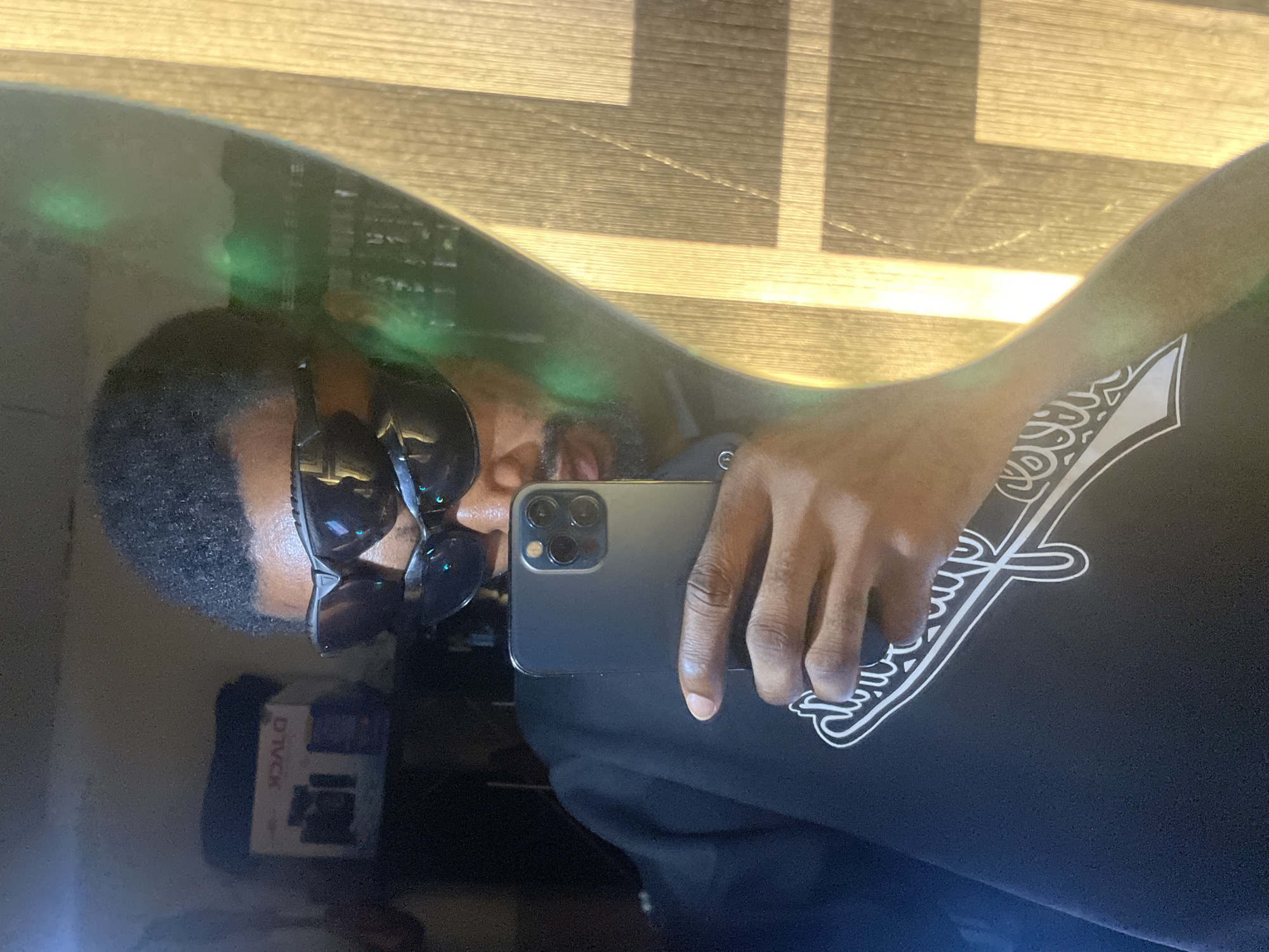 A male individual is captured in a mirror selfie taken in an indoor setting in Lokoja, Nigeria. The subject, with dark skin, short dark hair, and a beard, is wearing two stacked pairs of dark-lensed sunglasses that obscure his eyes. He is dressed in a black long-sleeved shirt featuring a partial white graphic design on the chest. His right hand holds a dark smartphone, identified by its triple camera lens array, positioned to capture the reflection. The background reflected in the mirror includes a dark interior space on the left, where a white box with reversed text and graphics is visible on a surface. To the right, a wooden surface with a vertical grain pattern is illuminated by a strong light source, casting defined highlights and shadows. Green lens flares are present on the mirror's surface, notably on the right side.