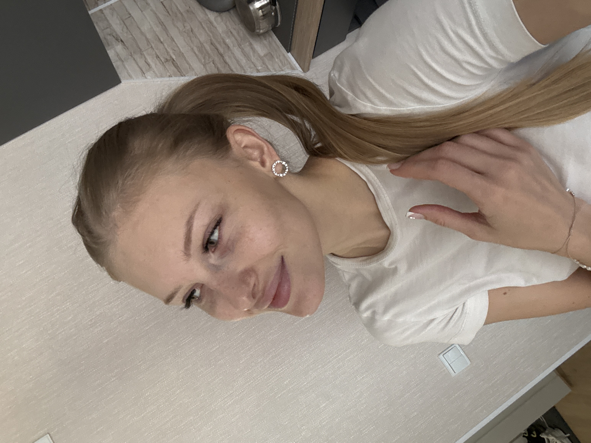 A young woman with blonde hair in a ponytail, wearing a white t-shirt and circular earrings, looks to her left while resting her hand on her chest. She is indoors, in front of a textured wall, with a glimpse of a tiled wall and a countertop in the background. The environment suggests a residential or personal space. The lighting is natural, casting soft shadows.