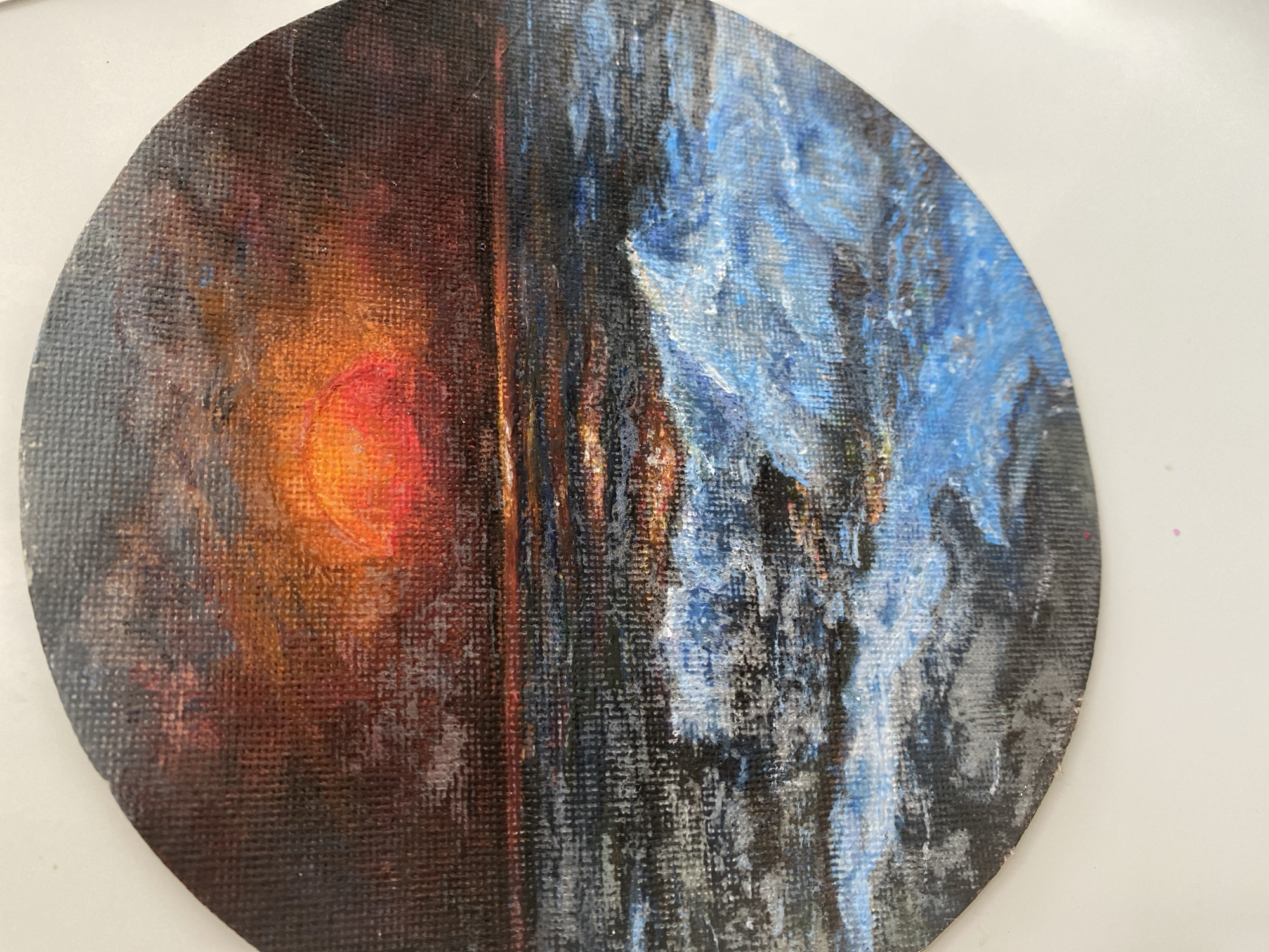 The image is a circular painting of a seascape at sunset. The sun, a vibrant orange orb, is low on the horizon, casting a warm glow across the sky and reflecting on the water. Dark, textured clouds are painted in shades of red, orange, and dark blue, creating a dramatic sky. The ocean is depicted with choppy waves in shades of deep blue, gray, and white foam. There are no people or man-made objects visible in the painting. The painting is presented on a white background. There is no information in the image to suggest its location in the city of Sokhumi, Abkhazia.