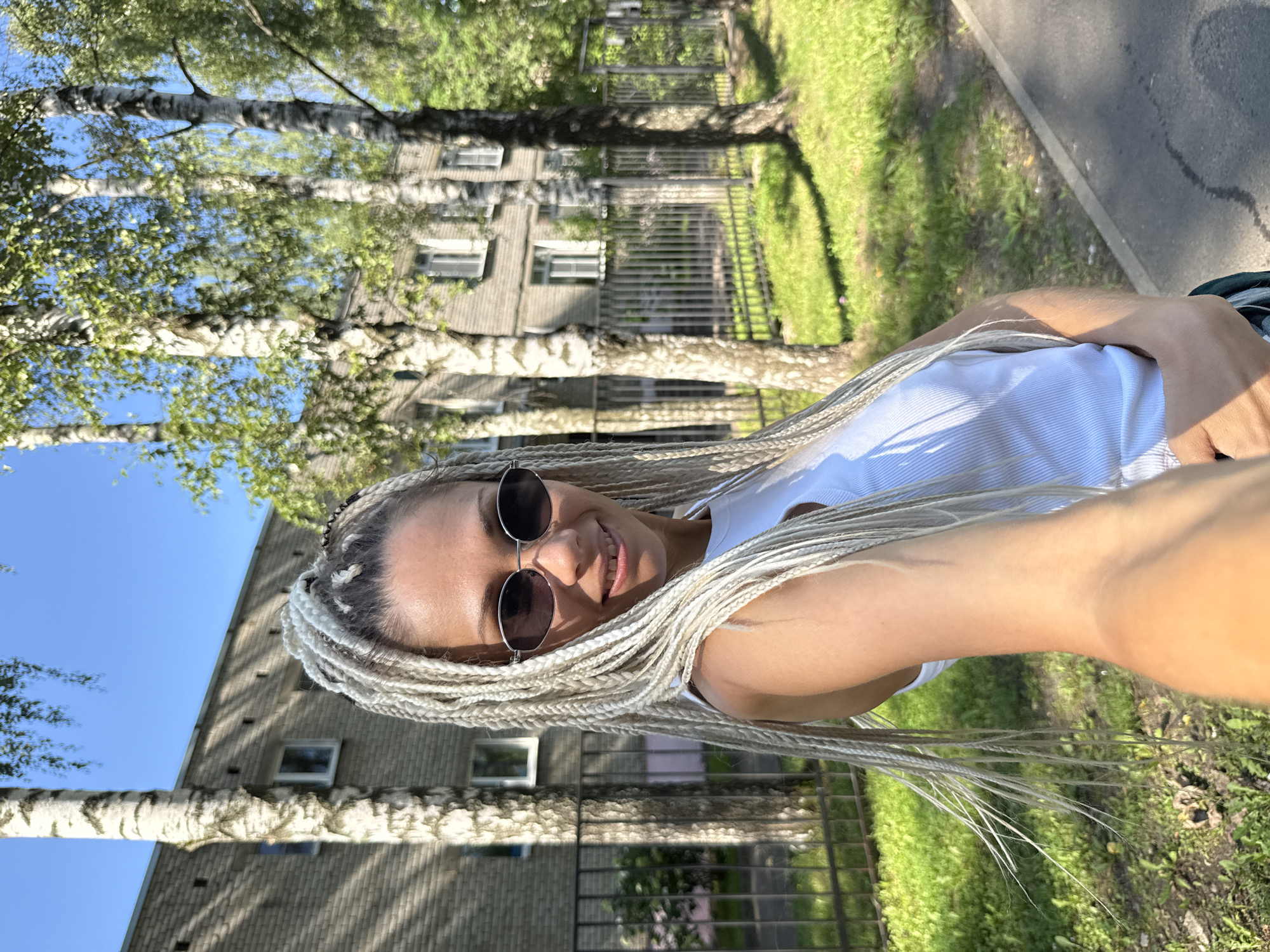 The image is a selfie taken outdoors on a sunny day. The subject is a woman with blonde braided hair, wearing a white tank top and sunglasses. She is smiling at the camera, and her hair is a prominent feature, cascading around her shoulders and face.
The background shows a building with multiple windows, likely an apartment building, and several trees with green leaves. A paved pathway runs along the right side of the image, bordered by grass. A metal fence is visible in front of the building. The scene appears to be a residential area, possibly a park or a street with trees and buildings.
The lighting suggests it is daytime, with bright sunlight casting shadows. The overall mood is cheerful and relaxed, as indicated by the woman's smile. The location context is Rusanovo, Russia. There is no visible text in the image.