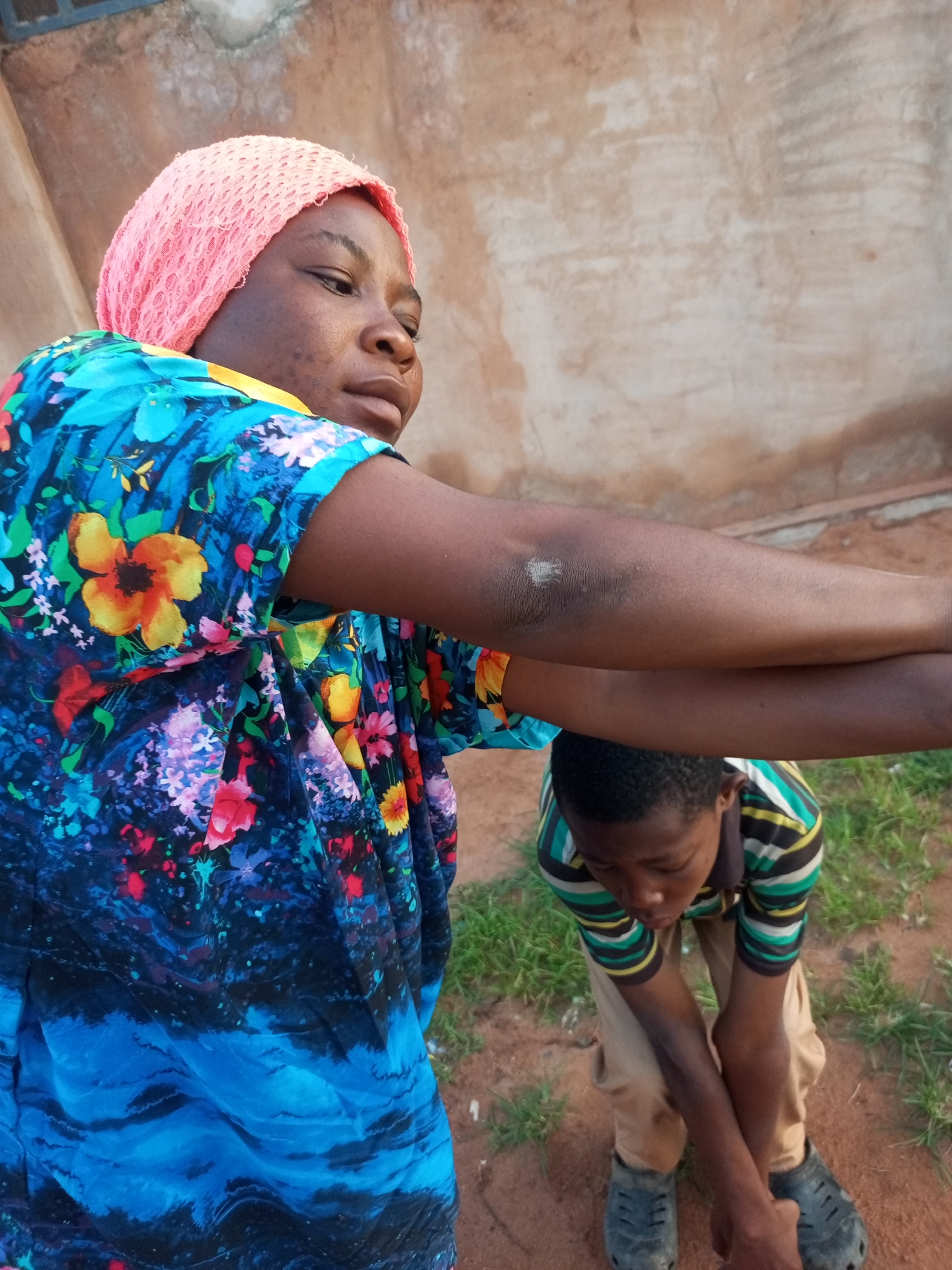 A woman wearing a pink headscarf and a blue, floral-patterned dress is positioned in the foreground, facing to the right. Her arms are extended forward, with her left arm crossed over her right. Her gaze is directed downwards and to the right. Behind her, a boy in a striped shirt and khaki pants is bending forward, his arms clasped in front of him. He is looking down. The background consists of a rough, light-brown wall and patches of dry grass and dirt. The setting appears to be an outdoor area in Tempure, Nigeria, possibly a residential courtyard or street.