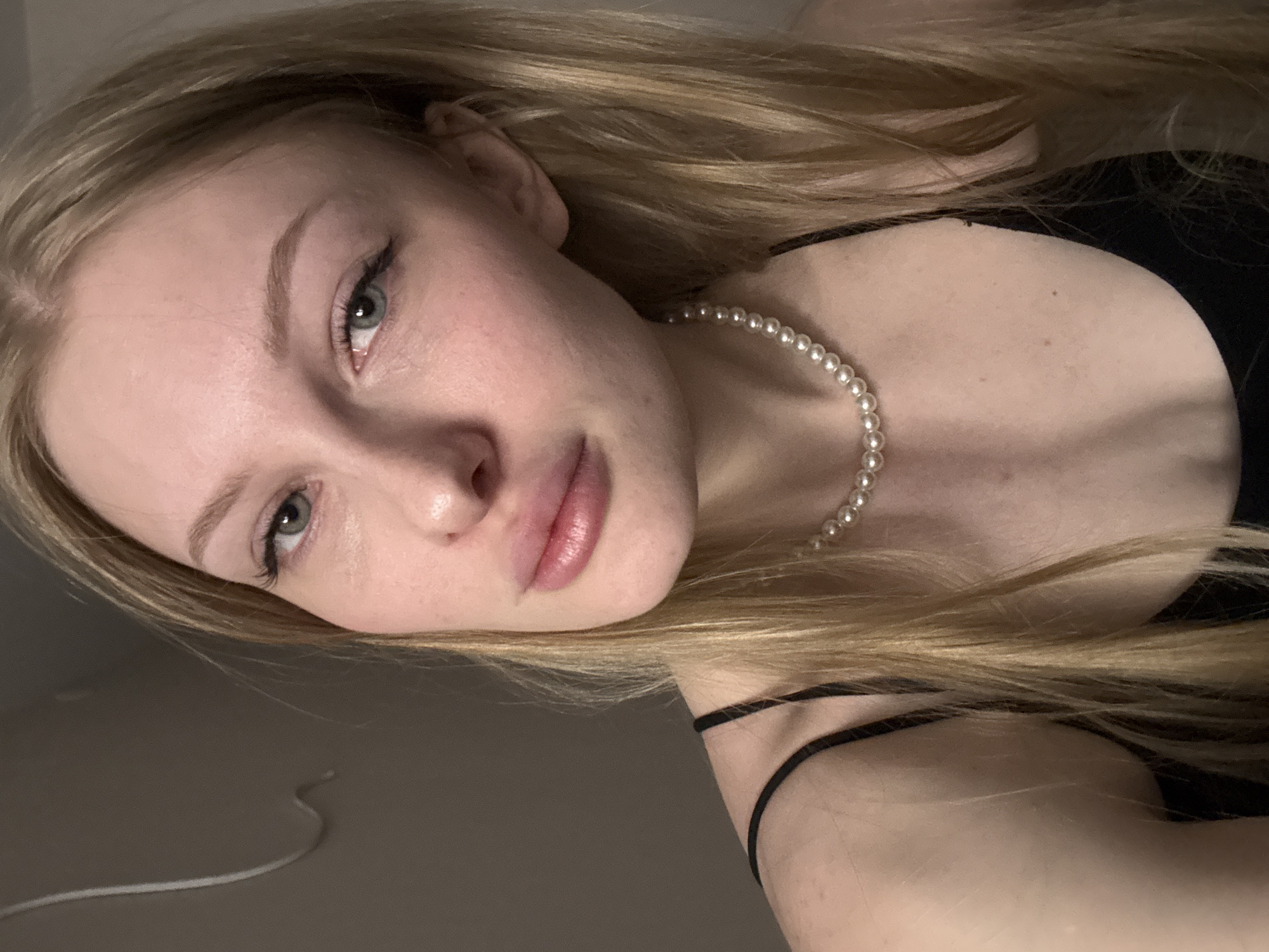 The image is a close-up, high-angle selfie of a young woman with fair skin and long, blonde hair. She is wearing a black tank top and a pearl necklace. Her makeup includes eyeliner and mascara. The background is a plain, dark gray wall with a white cord hanging loosely. The lighting is dim and appears to be from a single source, casting soft shadows. The image is a portrait and does not depict any specific location in the city of Perm, Russia.
