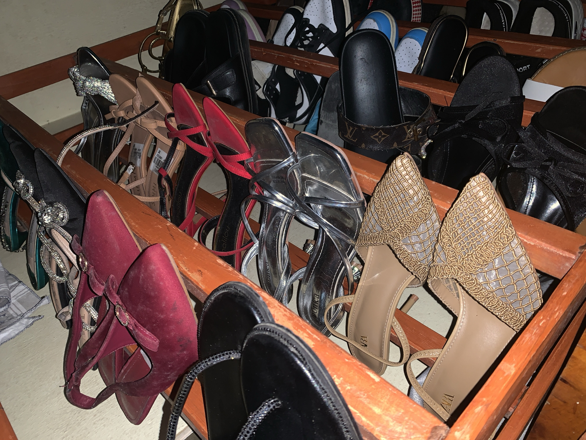 A wooden shoe rack is positioned indoors, holding an assortment of women's footwear. The rack contains high heels in red, silver, black, and gold tones. Some heels are decorated with bows or straps. Several pairs of black sandals and a pair of white sneakers are also visible. The background is nondescript, suggesting an interior space in a residential setting. There are no people visible in the scene. The location cannot be verified from the image.