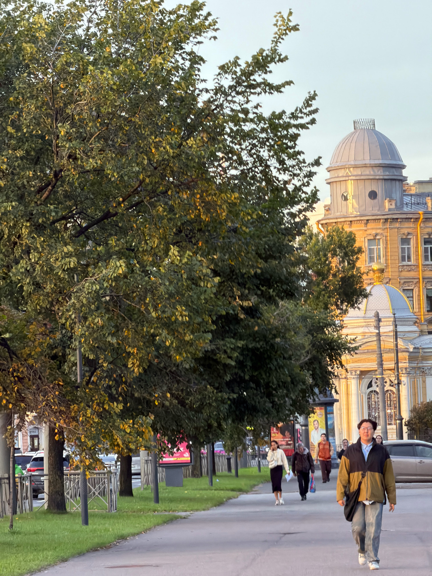 The image depicts a street scene in Saint Petersburg, Russia, likely during the daytime. In the foreground, a paved walkway runs alongside a grassy area lined with trees. Several people are walking on the walkway. The person closest to the camera is a young man wearing glasses, a light blue collared shirt, a two-toned jacket (khaki and dark brown), and light-wash jeans. He is carrying a black bag slung across his chest. Further down the walkway, other individuals are walking in the same direction. A woman in a white top and black skirt, a man in a dark jacket carrying a blue bag, and another person in an orange jacket are visible. In the background, there are buildings with classical architecture, including domes, and a street with cars. The trees lining the walkway have green leaves, with some showing hints of yellow, suggesting it might be early autumn. The overall atmosphere is that of a serene urban environment.