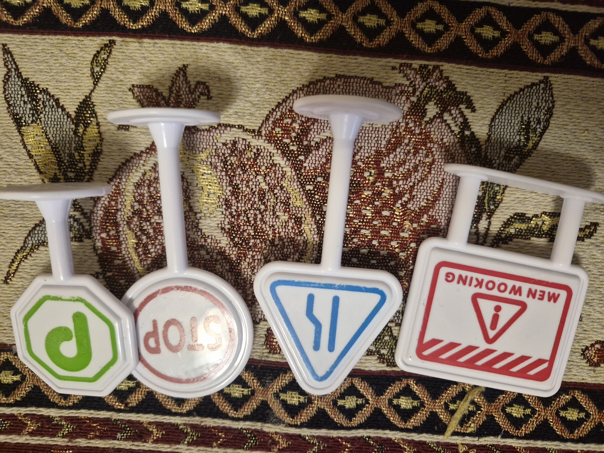 The image shows four small, white plastic toy road signs laid out on a patterned fabric surface, likely a tablecloth or tapestry, with shades of beige, brown, gold, and red. The lighting suggests an indoor setting with artificial light casting shadows from the signs.
From left to right, the signs are:
1. An octagonal sign with a white background, a green border, and a green stylized character that looks like a capital 'D'.
2. A circular sign with a white background, a red border, and the word "STOP" printed in red, but it is upside down and mirrored, appearing as "dOLS".
3. A triangular sign with a white background, a blue border, and a blue symbol depicting two vertical lines, one straight and one slightly curved, possibly indicating a road narrowing or merging, also appearing mirrored.
4. A rectangular sign with a white background, a red border, and red diagonal stripes at the bottom. It features a red triangular warning symbol with an exclamation mark, and above it, the misspelled phrase "MEN WOOKING" printed in red, which is upside down and mirrored. This is likely intended to be "MEN WORKING".
The signs appear to be children's toys due to their small scale, plastic material, and the incorrect orientation and spelling of the text. No activity or event is taking place, only the static display of these objects in an indoor environment in Yerevan, Armenia.