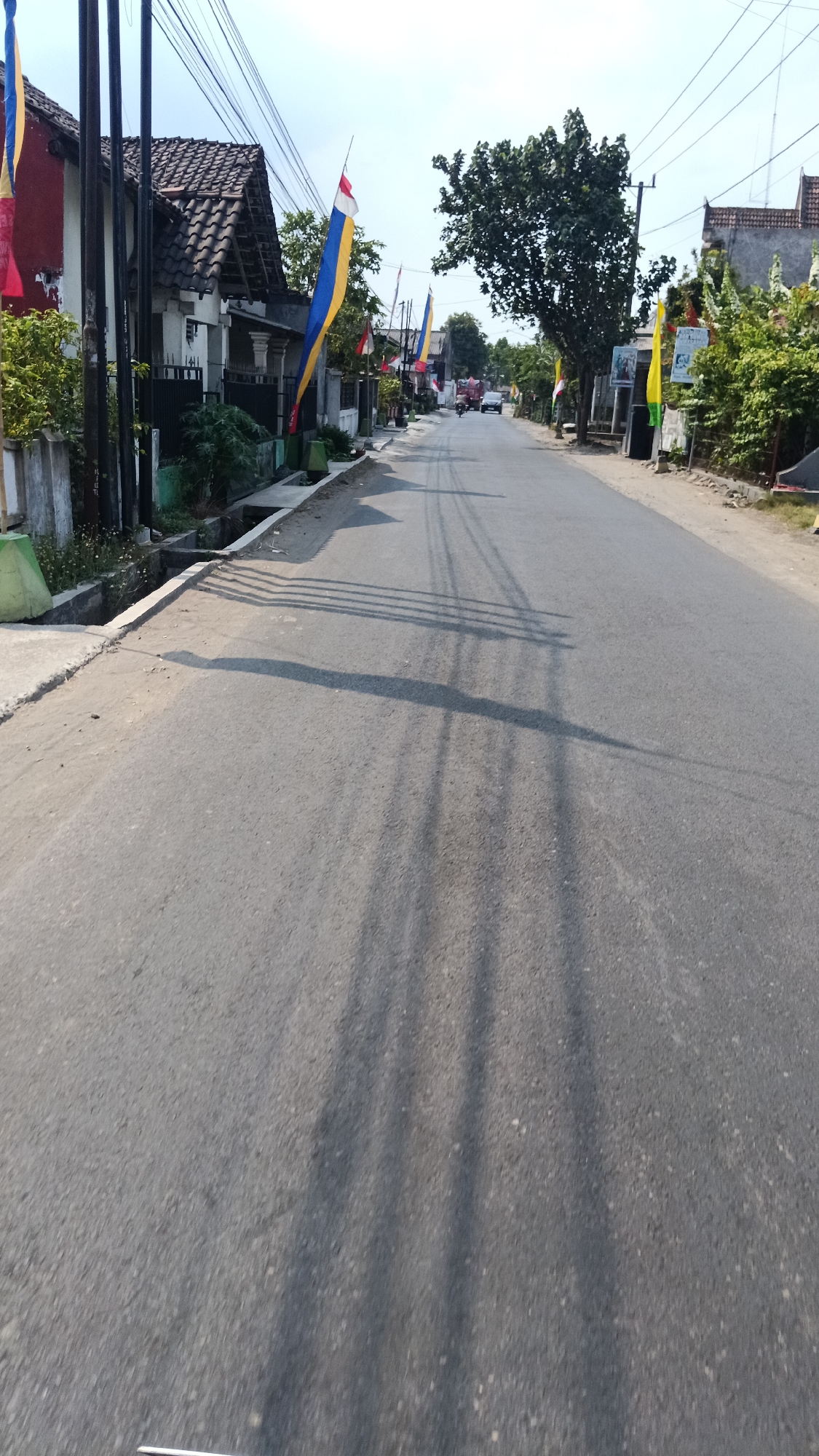 This is a street-level, eye-level shot of a paved road in Cendono, Indonesia, likely taken during the daytime. The road runs into the distance, flanked by houses on the left and right. Several tall, slender poles, possibly for utility lines, are visible on the left side of the road, casting long shadows across the asphalt.
Along the left side of the road, a row of houses with tiled roofs is visible, with some greenery and small plants growing near the roadside. Several tall, colorful flags, red, white, and blue with yellow accents, are planted along the roadside, suggesting a festive occasion or national celebration.
On the right side of the road, more houses are visible, as well as trees and dense foliage. The road appears to be in a residential area. In the distance, a few vehicles, including a red truck and other cars, are traveling on the road. The sky is mostly clear with some visible clouds. The overall impression is of a quiet, everyday scene in a small Indonesian town.
