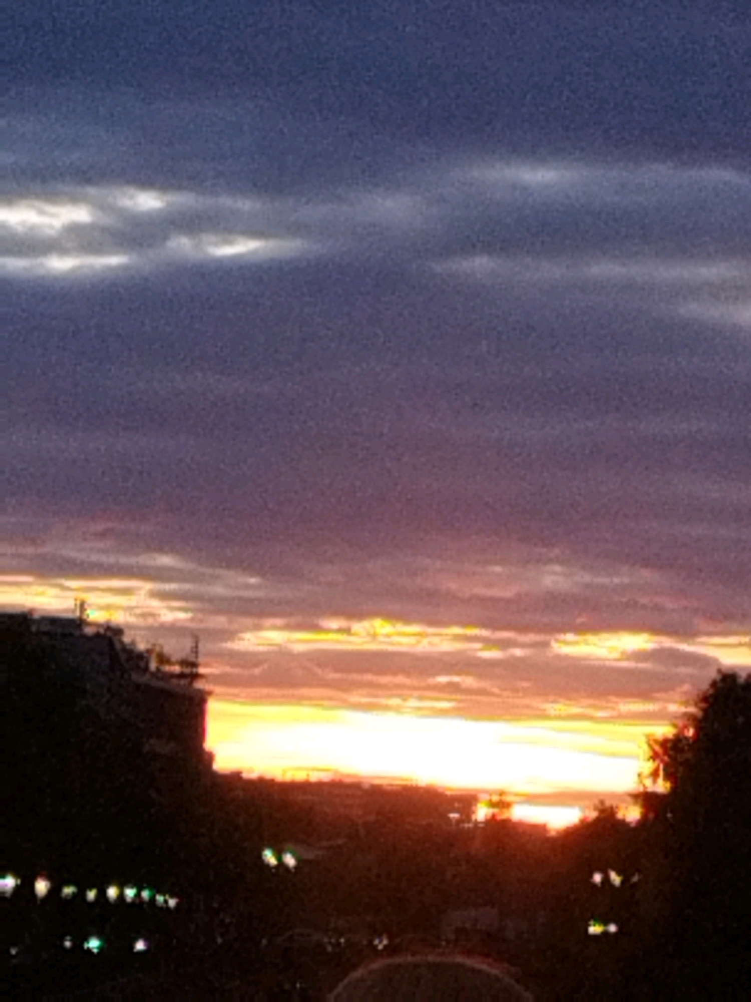 The image depicts a sunset over Bucharest, Romania, viewed from a distance. The sky is filled with layers of clouds, transitioning from a deep blue at the top to shades of purple and pink, and finally to bright yellow and orange hues near the horizon, indicating the sun is setting. Silhouetted against the colorful sky are the dark shapes of buildings and trees. In the foreground, the outlines of some structures and scattered lights are visible, suggesting an urban environment. The overall impression is a serene, twilight cityscape.