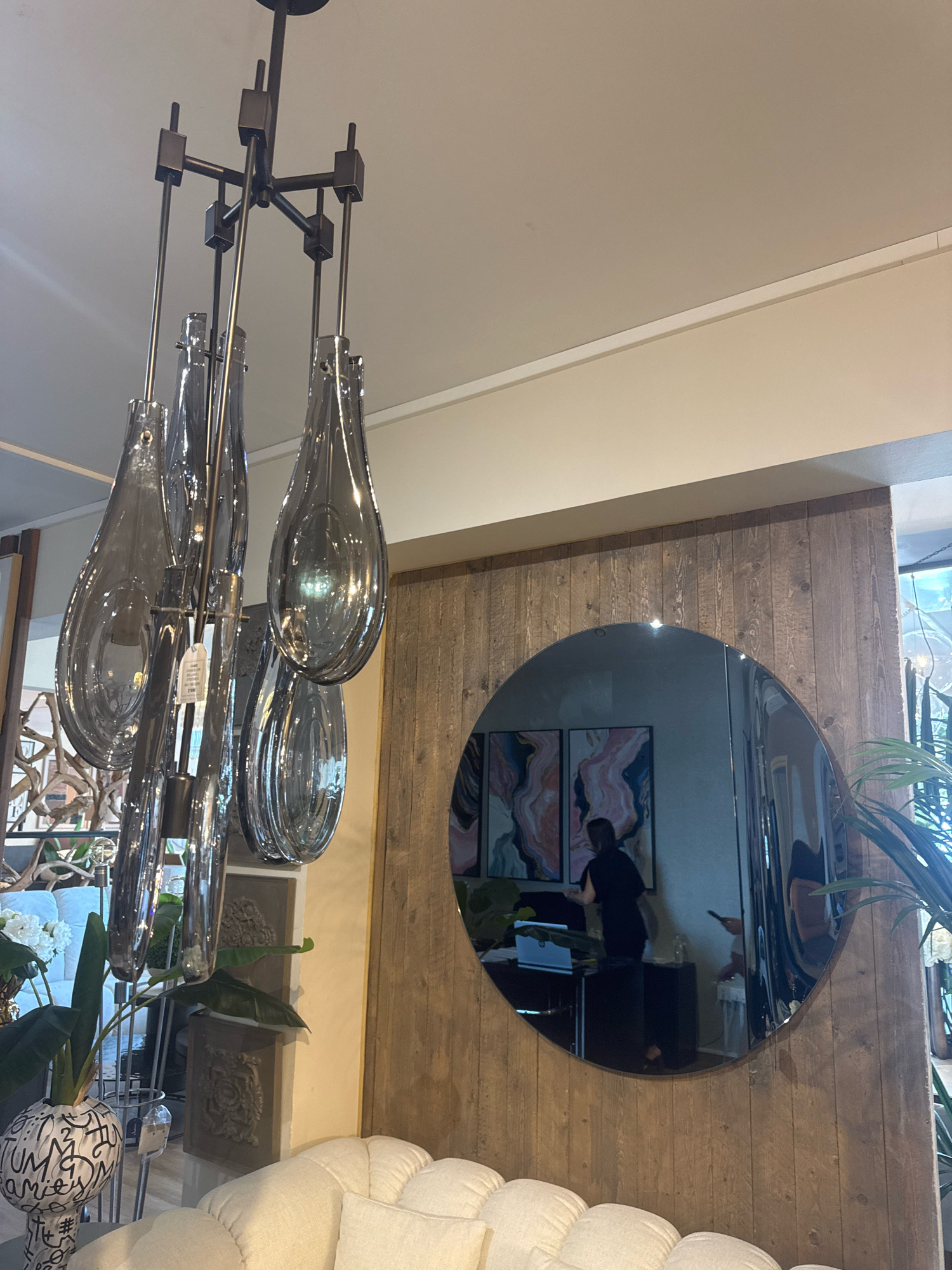 A dimly lit interior space features a modern chandelier with teardrop-shaped grey glass elements hanging from the ceiling. A large, circular, highly reflective mirror is mounted on a wall clad in rustic wooden planks. The mirror reflects two abstract framed artworks with swirling pink, blue, and white patterns, and a person dressed in dark clothing standing in front of a desk. In the foreground, a plush white sofa is partially visible, with a white ceramic vase decorated with black graffiti-style lettering to its left. A large-leafed plant is positioned behind the vase. The environment suggests a showroom or a well-decorated living space. The location is identified as Agios Athanasios Municipality, Cyprus, based on its context within a larger dataset related to this region.