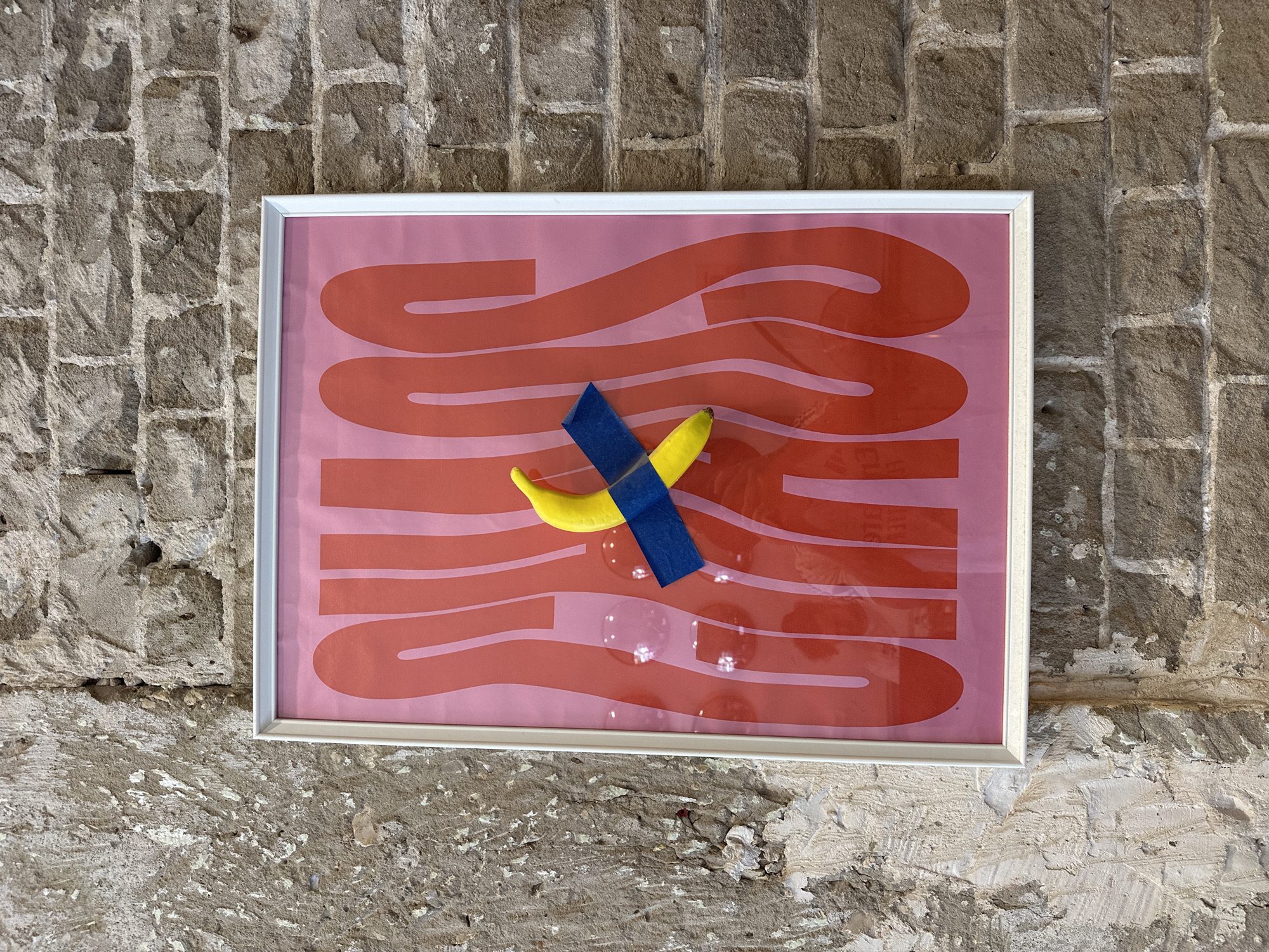 A framed piece of art hangs on a rough, light-colored brick wall. The frame is white. Inside the frame is a pink and red abstract design. Affixed to the front of the framed art is a yellow banana held in place by blue tape. There are some water droplets below the banana. The location is assumed to be somewhere in the city of Tel-Aviv, Israel, but there is no further evidence of the precise location.
