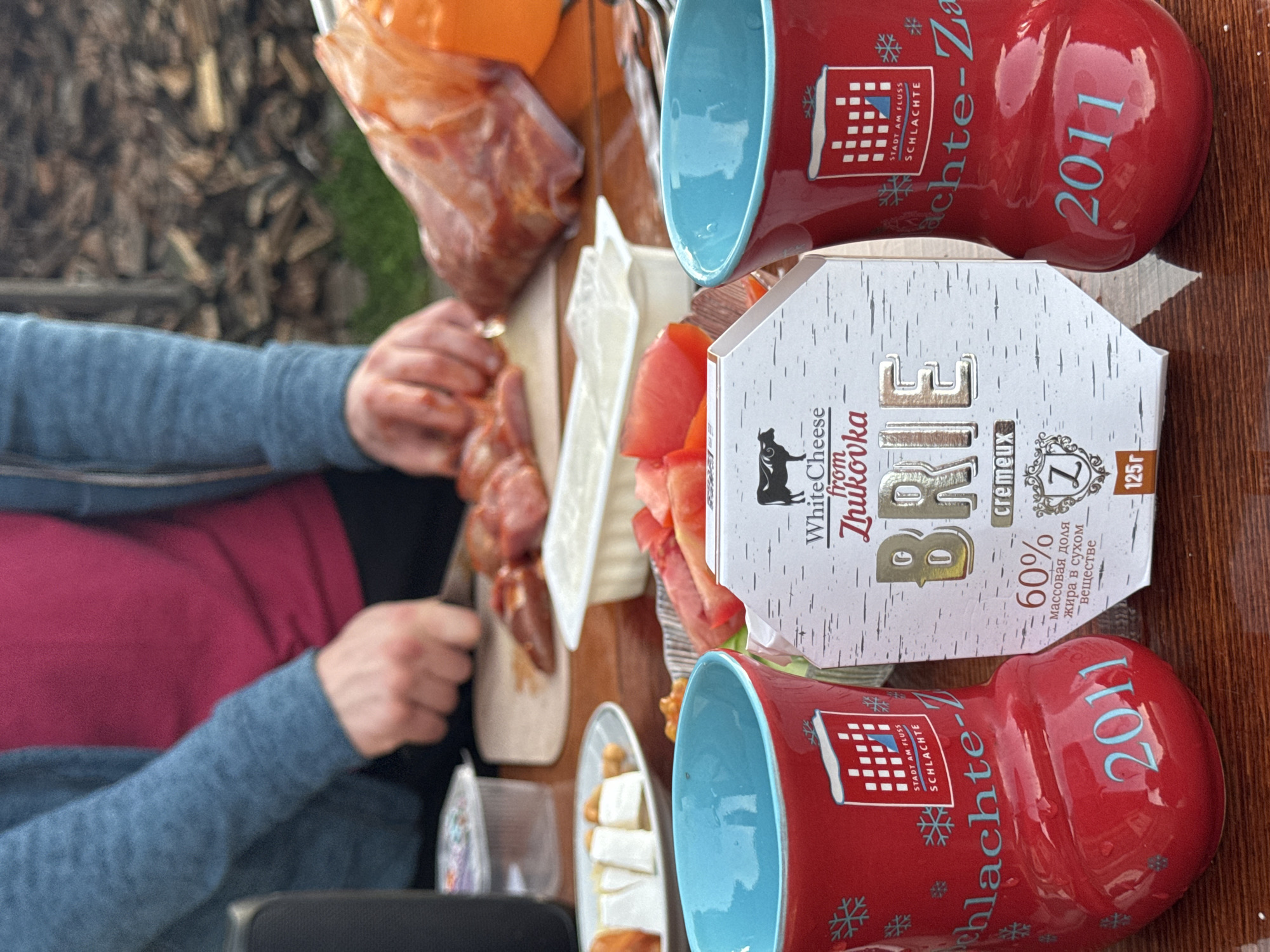 The image captures an outdoor scene with a wooden table as the primary surface. A person, partially visible from the torso down, is in the process of slicing meat on a wooden cutting board. Their attire includes a blue sweater over a pink shirt.
Scattered on the table are various food items. In the foreground, two red mugs with blue interiors are prominent, each featuring a logo and the year "2011" in blue lettering. One mug has snowflake designs. Between the mugs, a box of "BRIE" cheese, with text indicating "White Cheese from Zhukovka," is positioned. The cheese box has a stylized birch bark design and mentions "60% жира в сухом веществе" (60% fat in dry matter) and "125г" (125g).
To the left of the cheese box, a plate holds slices of cheese and nuts. Further back, a white container holds a creamy substance, possibly yogurt or sour cream, and a platter displays sliced tomatoes and lettuce. A large bag of red meat is visible on the right side of the table, partially obscuring an orange object. In the background, a stack of firewood suggests a rustic or rural setting. The overall ambiance is informal, possibly a picnic or outdoor meal preparation. There is no information in the image that specifies a location within the city of Maksimovo, Russia.