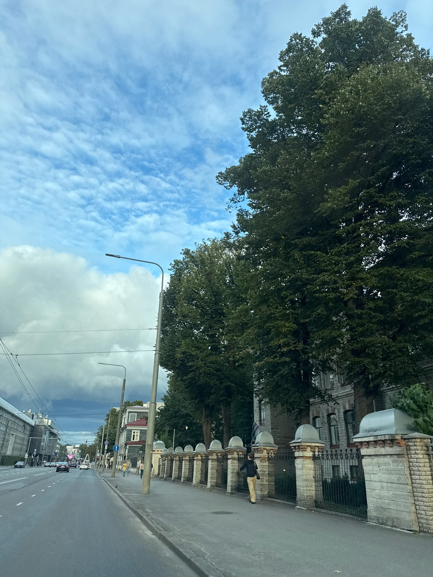 The image depicts a street scene in Tallinn, Estonia, likely on a cloudy day. A wide asphalt road dominates the foreground, with lane markings visible. To the right of the road is a sidewalk lined with a decorative stone fence featuring arched openings and capped pillars. Behind the fence, a large, mature tree with dense green foliage stands tall, partially obscuring a grey, multi-story building. A person wearing a dark jacket and light-colored trousers is walking away from the viewer on the sidewalk. Further down the street, several other buildings and vehicles are visible, including cars and what appear to be streetlights. The sky is a mixture of blue and white clouds, with darker clouds suggesting potential rain in the distance. The overall impression is of an urban environment with historical architectural elements and lush greenery.