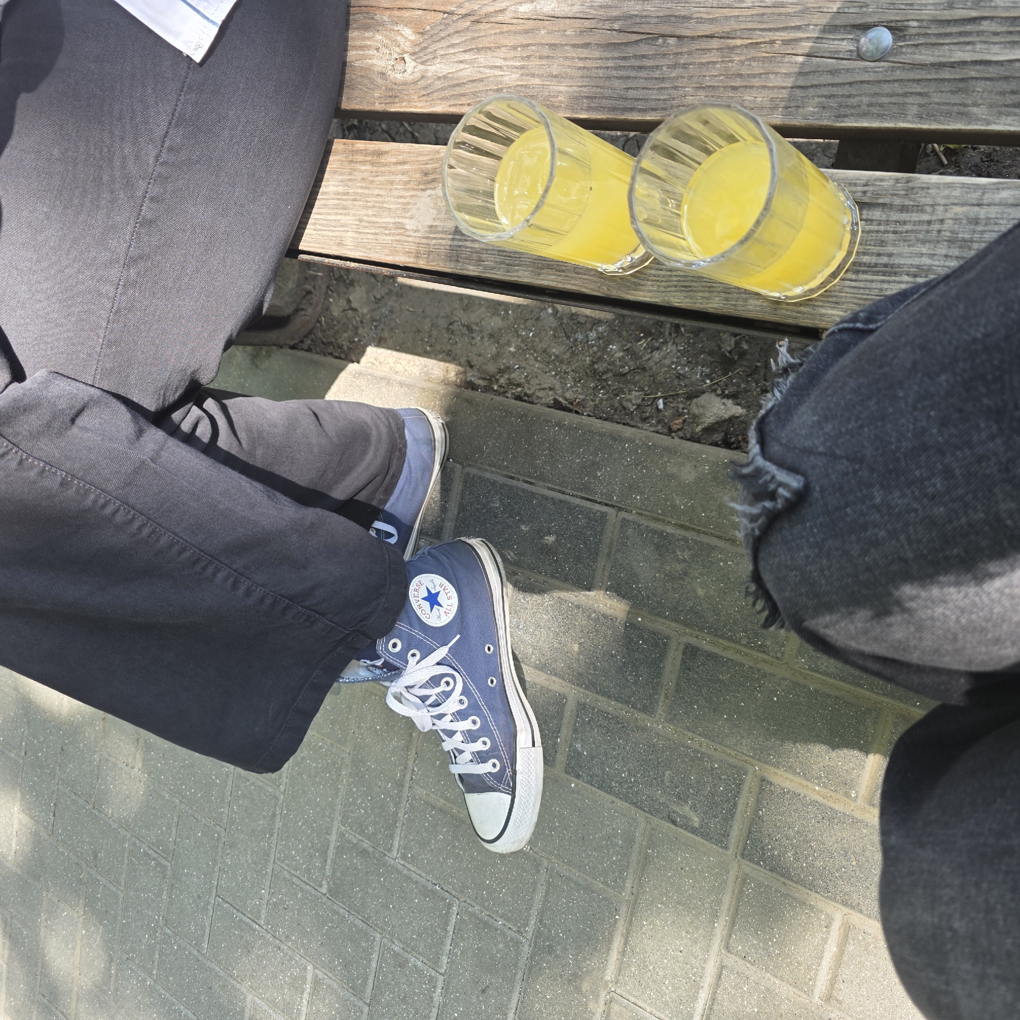 Two individuals are seated on a wooden bench outdoors. The person on the left is wearing blue Converse sneakers and dark pants. The person on the right is wearing dark pants and a white shirt sleeve is visible. In front of them on the bench are two glasses filled with a yellow beverage, possibly juice. The ground is paved with rectangular gray stones. The scene appears to be a casual setting, possibly a park or an outdoor seating area.