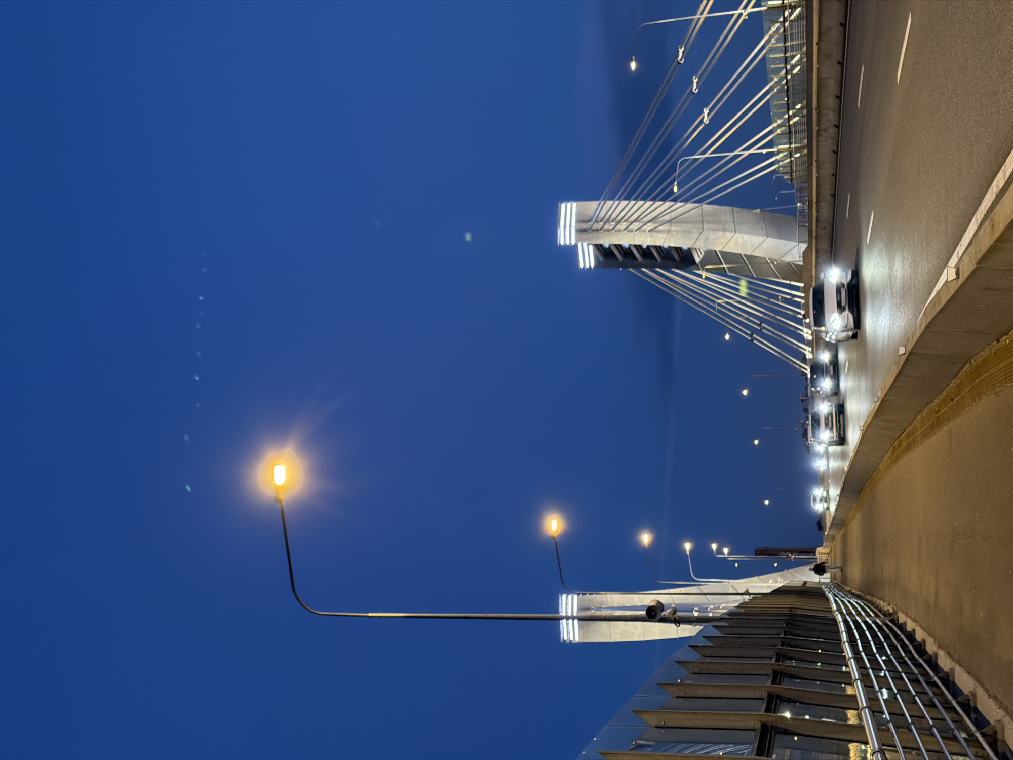 The image depicts a view of the Big Obukhovsky Bridge in Saint Petersburg, Russia, at dusk. The sky is a deep blue, and the bridge's modern architecture, with its tall pylons and suspension cables, is illuminated. A sidewalk runs along the left side of the bridge, separated from the road by a metal railing. Several streetlights are visible, casting a warm glow on the path. On the right side of the image, cars are driving on the road, their headlights and taillights creating streaks of light. One person is walking on the sidewalk in the distance. The overall scene conveys a sense of urban infrastructure and nighttime activity.