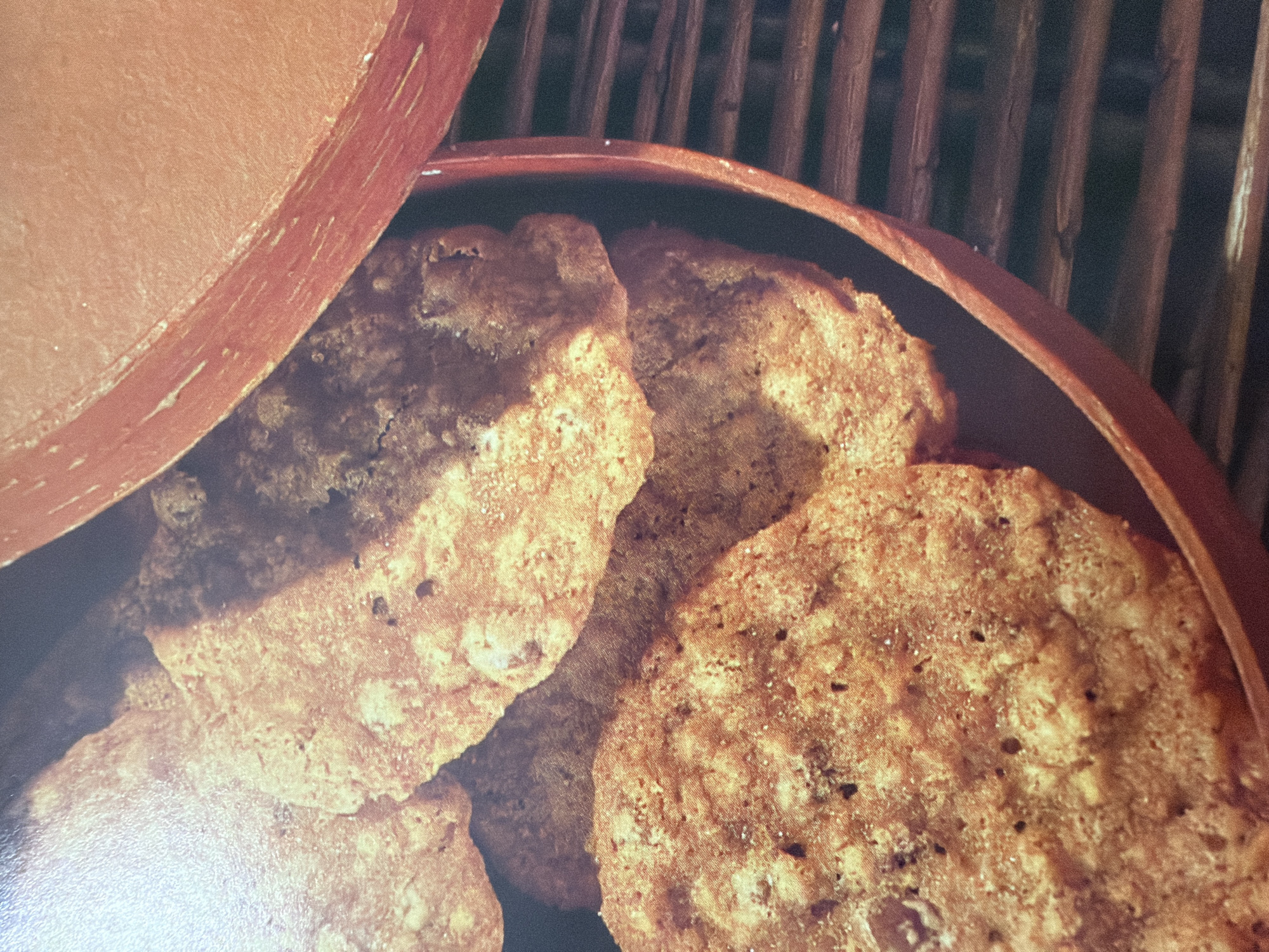 This is a close-up, overhead shot of oatmeal cookies in a red bowl. The cookies are golden brown and appear to be homemade with visible oats and possibly chocolate chips. The bowl is partially filled with cookies, and a portion of the bowl's rim is visible, along with some woven material underneath. The lighting creates shadows and highlights on the cookies, giving them a textured appearance. The image is set in an unknown location in Unknown, Unknown.