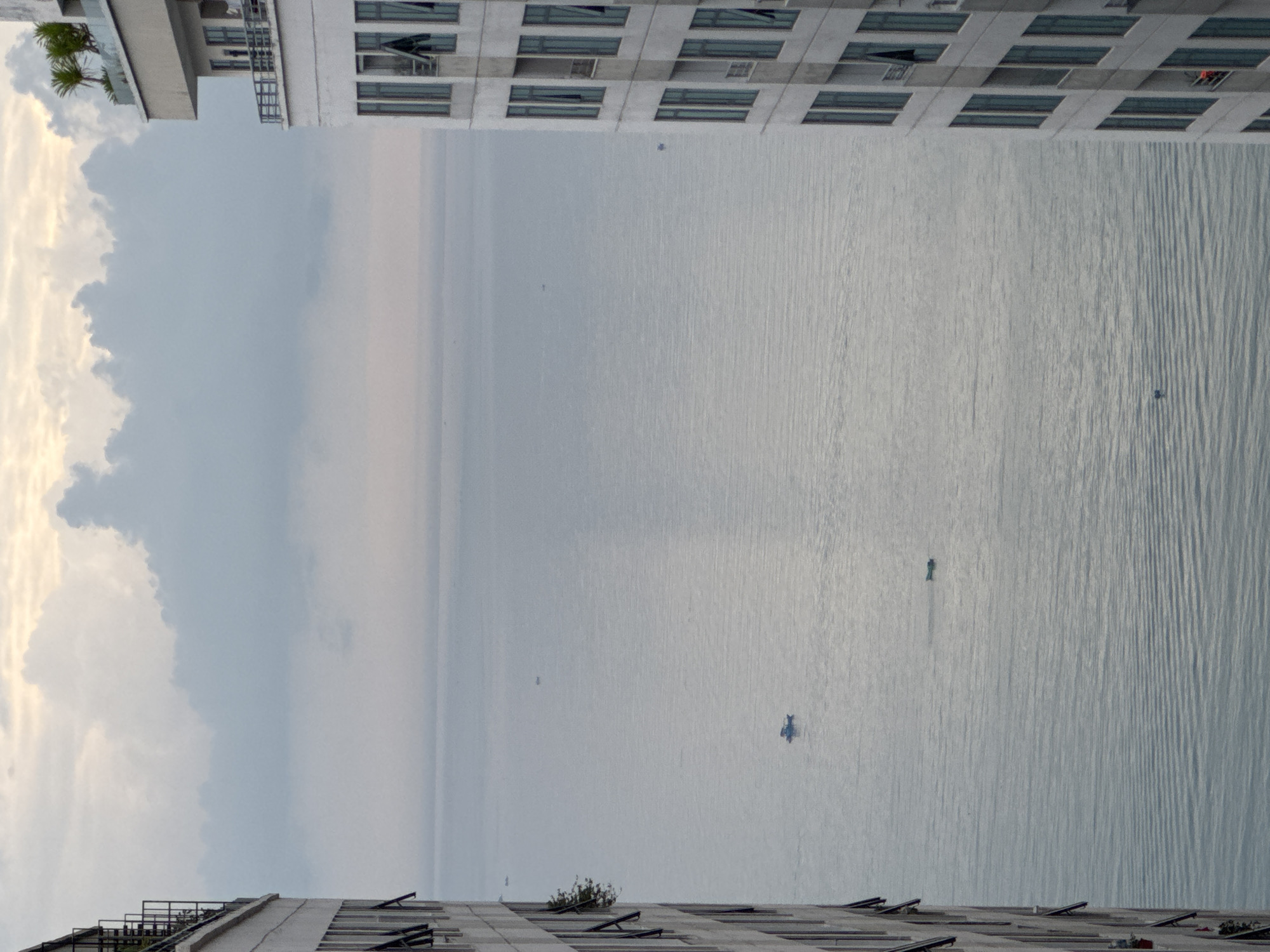 The image shows an expansive view of the sea from what appears to be a high vantage point in Bac Nha Trang Ward, Vietnam. Two tall buildings frame the scene, one on the top and another on the bottom. The sea is vast and stretches to the horizon under a cloudy sky. Several small boats are scattered across the water's surface.