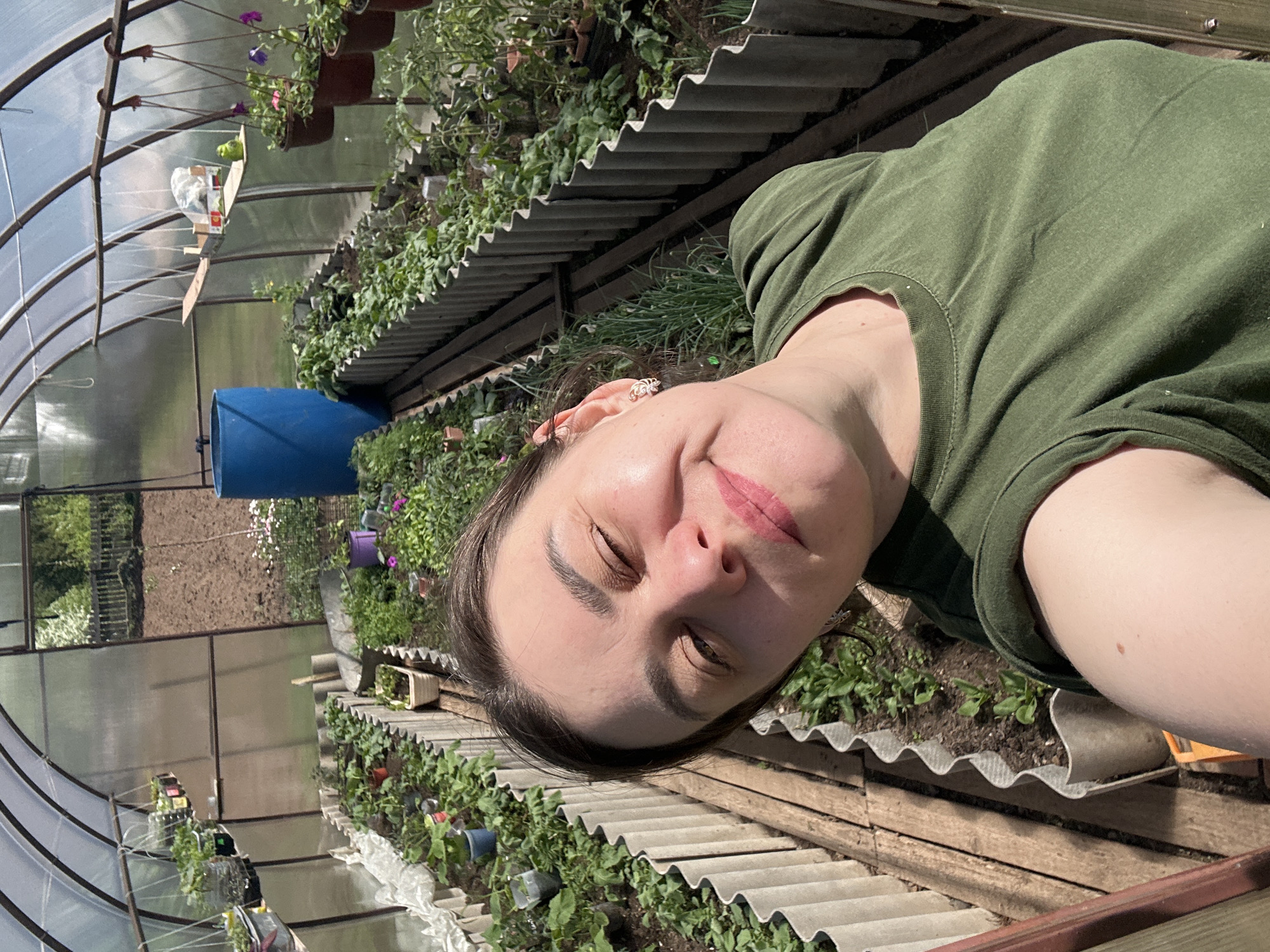 A woman wearing a green shirt is taking a selfie inside a greenhouse. Plants are growing in beds lined with corrugated material. A large blue barrel is visible in the background. The greenhouse is located in Фоки, Russia.